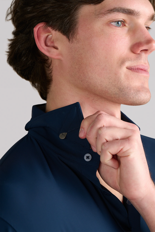 A light-skinned man with brown hair is seen in profile wearing the Twillory Event Polo Solid in Navy. He lifts its collar, revealing a small metal button underneath. The background is plain and light-colored.