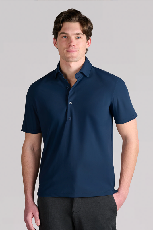 A man with short brown hair wears the Twillory Event Polo Solid in navy, made with performance stretch fabric, and dark pants. He stands facing forward, slightly smiling with his left hand in his pocket against a plain light gray background.