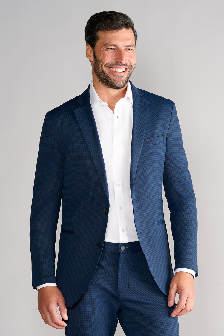 A man with short dark hair and a beard smiles against a light gray background, wearing Twillory's Performance Blazer in Navy Houndstooth over a white dress shirt, no tie, with his right hand relaxed at his side. A man with short dark hair and a beard smiles against a light gray background, wearing Twillory's Performance Blazer in Navy Houndstooth over a white dress shirt, no tie, with his right hand relaxed at his side.