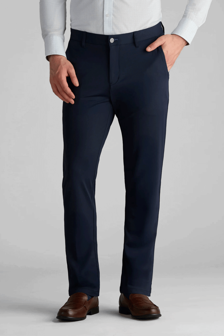A man wearing Twillory Performance Pants in navy blue, a light blue collared shirt, and brown leather dress shoes stands with one hand in his pocket against a plain light gray background. His head is not visible in the image. A man wearing Twillory Performance Pants in navy blue, a light blue collared shirt, and brown leather dress shoes stands with one hand in his pocket against a plain light gray background. His head is not visible in the image.