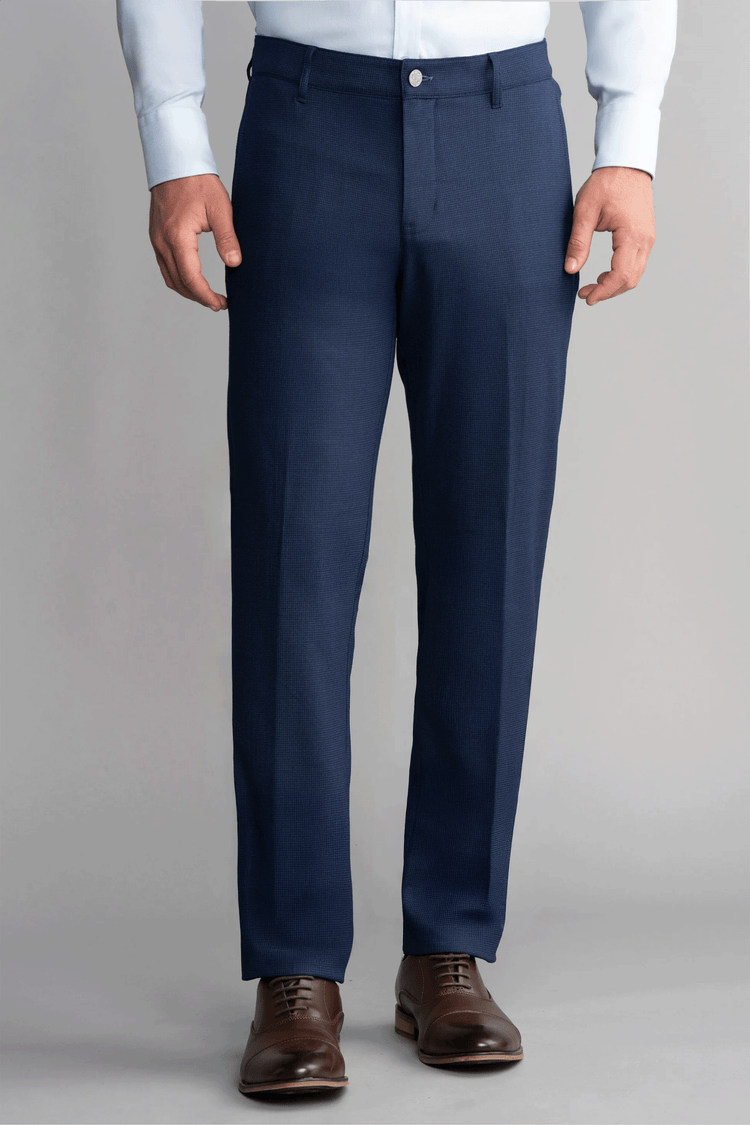 A person wears Twillory Hybrid Wool Pants in Navy Puppytooth, featuring a tailored fit and 4-way stretch, with a light blue dress shirt tucked in. Brown leather dress shoes complete the look against a plain light gray background. A person wears Twillory Hybrid Wool Pants in Navy Puppytooth, featuring a tailored fit and 4-way stretch, with a light blue dress shirt tucked in. Brown leather dress shoes complete the look against a plain light gray background.