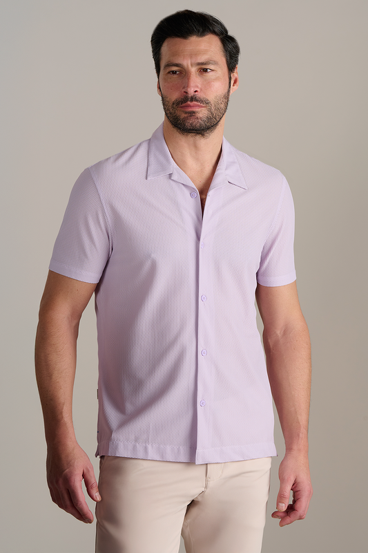 A man with dark hair and a beard wears the Twillory AIR Resort Shirt Solid in Lilac—made from cooling, machine-washable fabric—and light beige pants. He stands against a plain light gray background, looking slightly to the side.