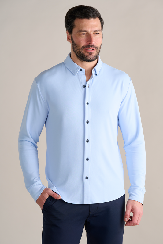 A man with short dark hair and a beard wears the Twillory Long Sleeve Button Down Polo Contrast in Powder Blue Navy, paired with dark pants. He stands against a beige background, left hand in his pocket and a neutral expression.