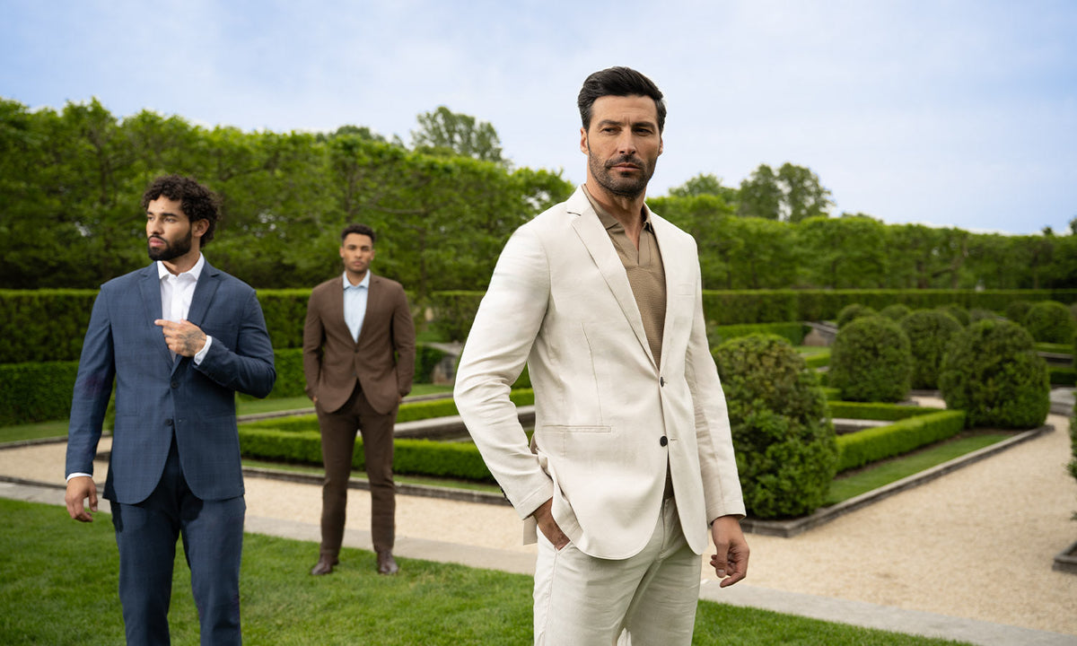Three men in stylish suits stand in a formal garden; the man in front wears a light suit, while the other two in blue and brown suits stand further behind amid manicured hedges on a bright day.
