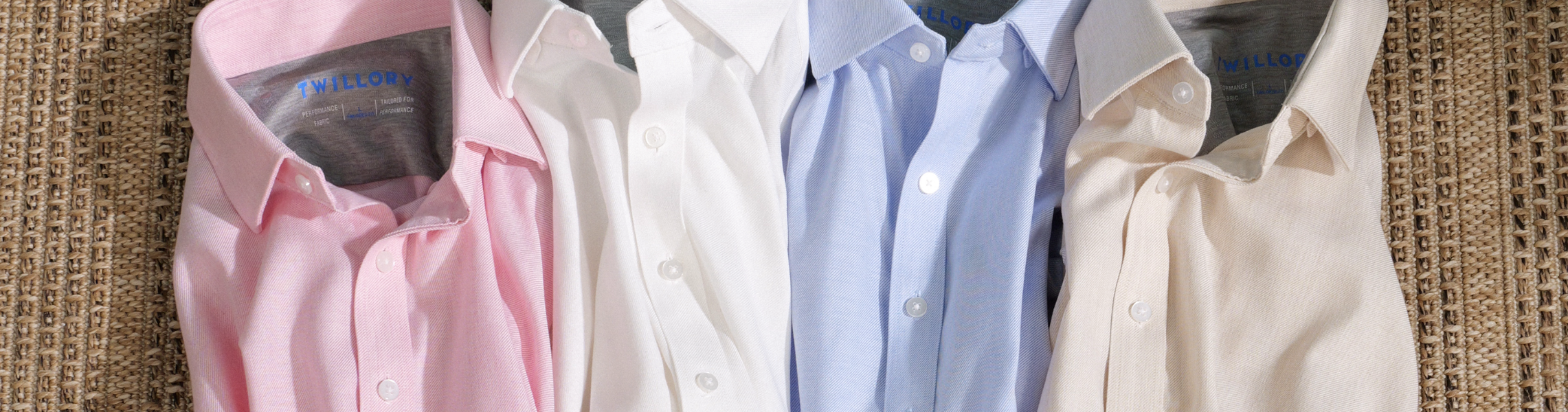 Four button-down shirts in pastel colors—pink, white, light blue, and beige—are neatly arranged side by side on a textured beige surface. The shirts all have collars and visible labels.