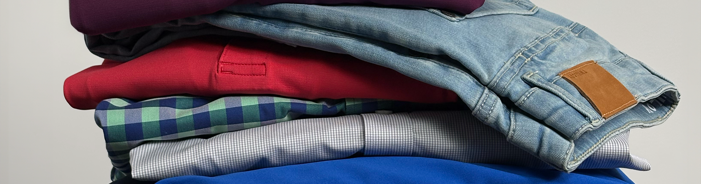 A neatly folded stack of clothes, including light blue denim jeans on top, followed by a red shirt, a green and blue plaid shirt, and other assorted shirts in various colors and patterns.