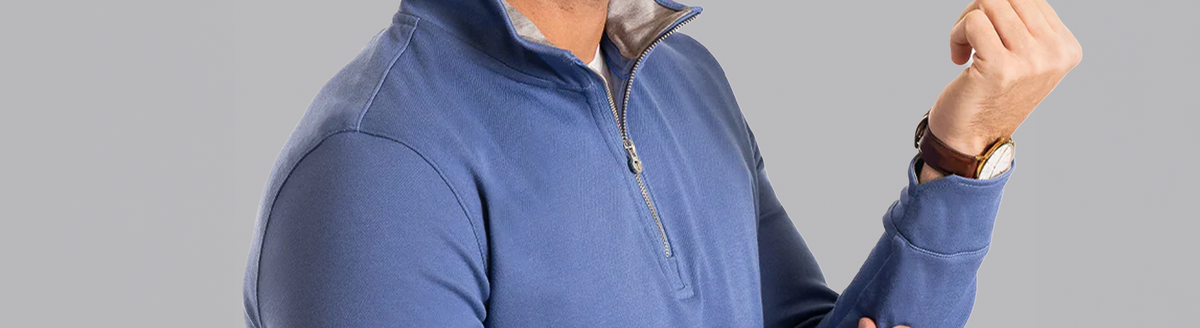A person wearing a blue quarter-zip pullover with a light gray collar lining, a white undershirt, and a brown leather watch, shown from the shoulders to the mouth, against a plain gray background.