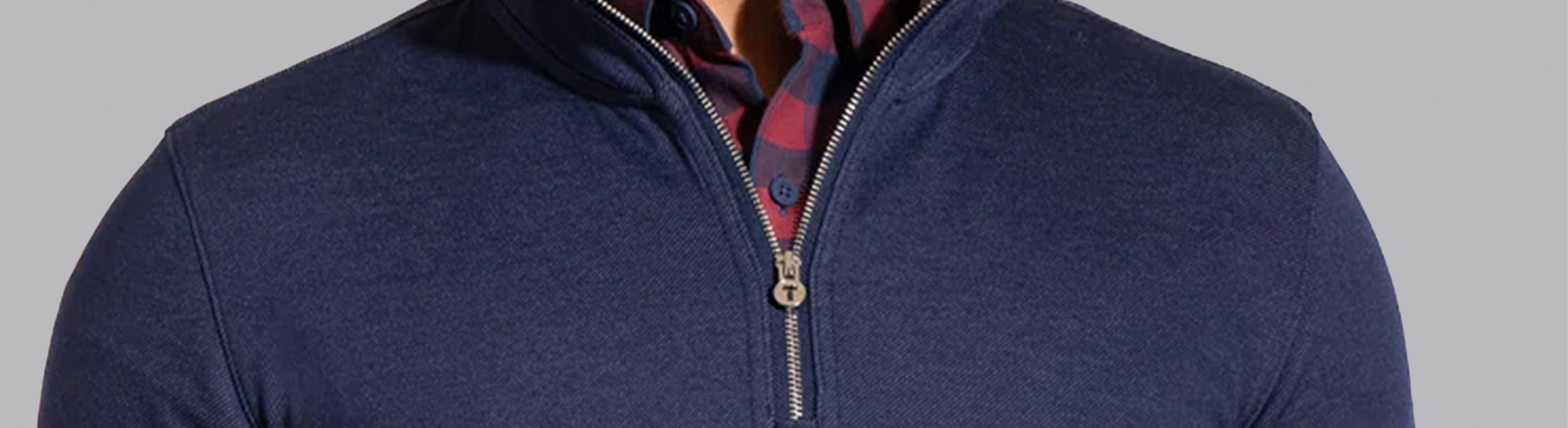 Close-up of a person wearing a navy half-zip sweater over a red and navy plaid button-down shirt, with the zipper partially open and the shirt collar visible above the sweater.