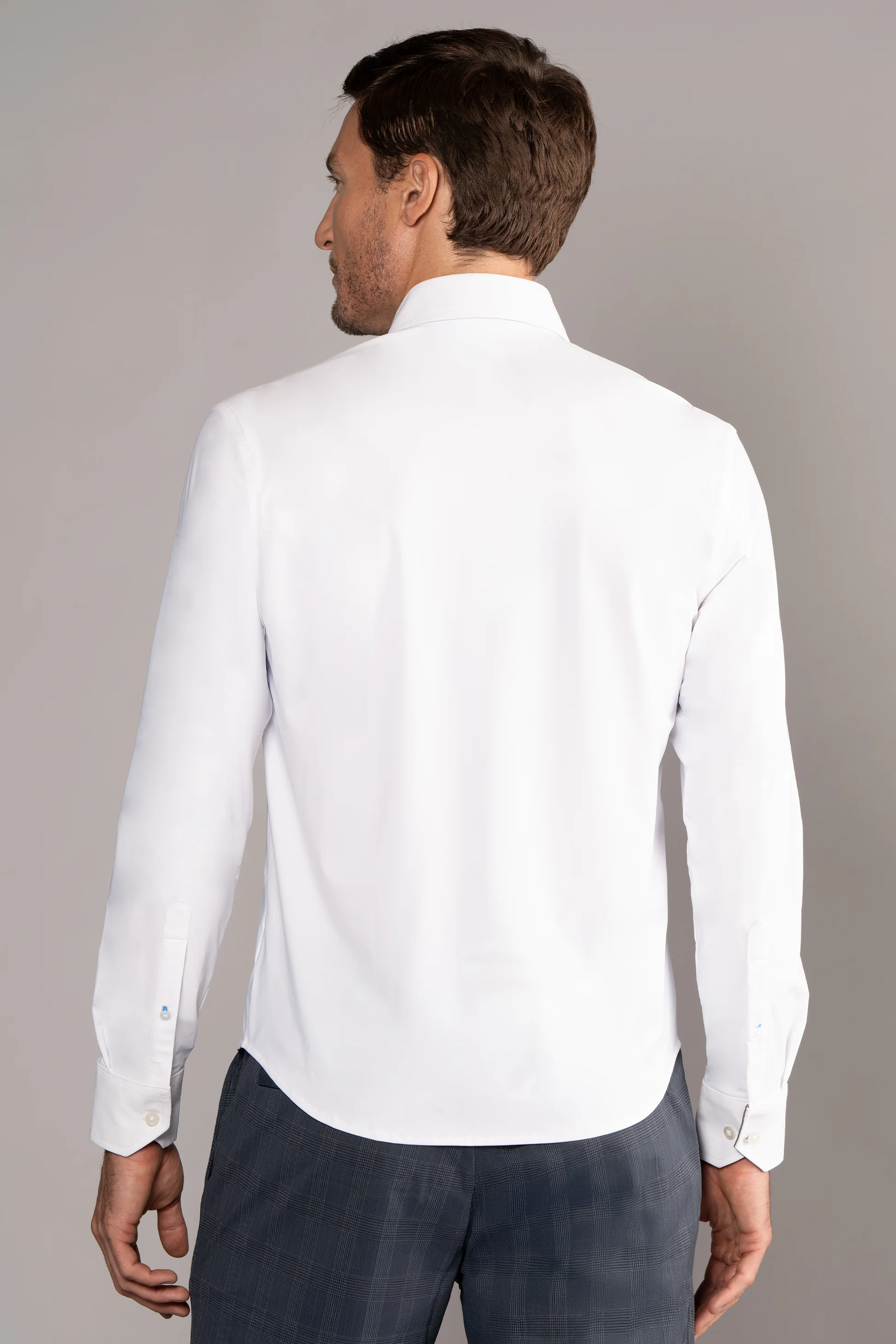 A man with short brown hair is seen from behind, wearing Twillory's Performance Linen Shirt in white and gray checkered dress pants. The fitted shirt features a crisp collar, buttoned cuffs, and wrinkle-free stretch fabric. The background is plain gray.