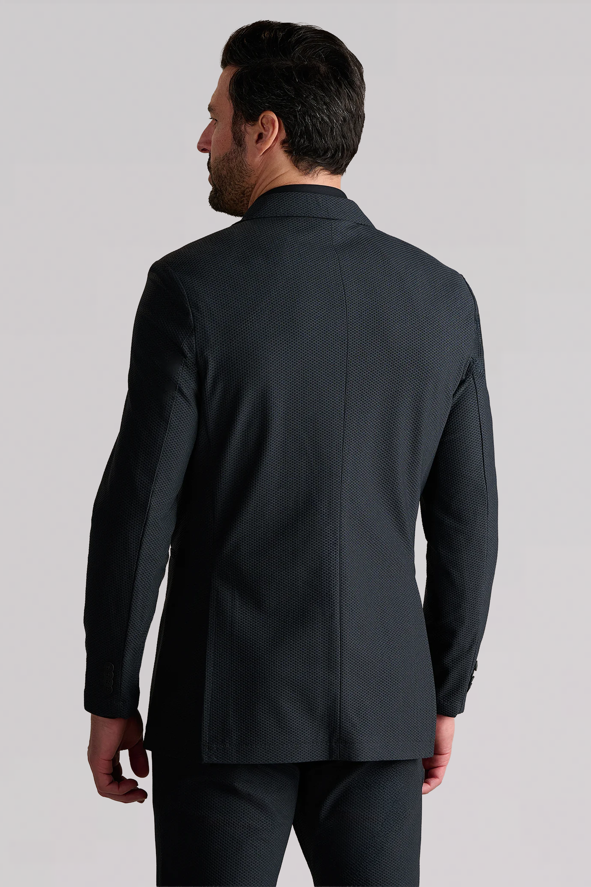 A man with dark hair is seen from behind wearing the Twillory AIR Blazer // Heather Charcoal, featuring a textured, breathable fabric and close fit. He stands against a light background with his left hand resting by his side.