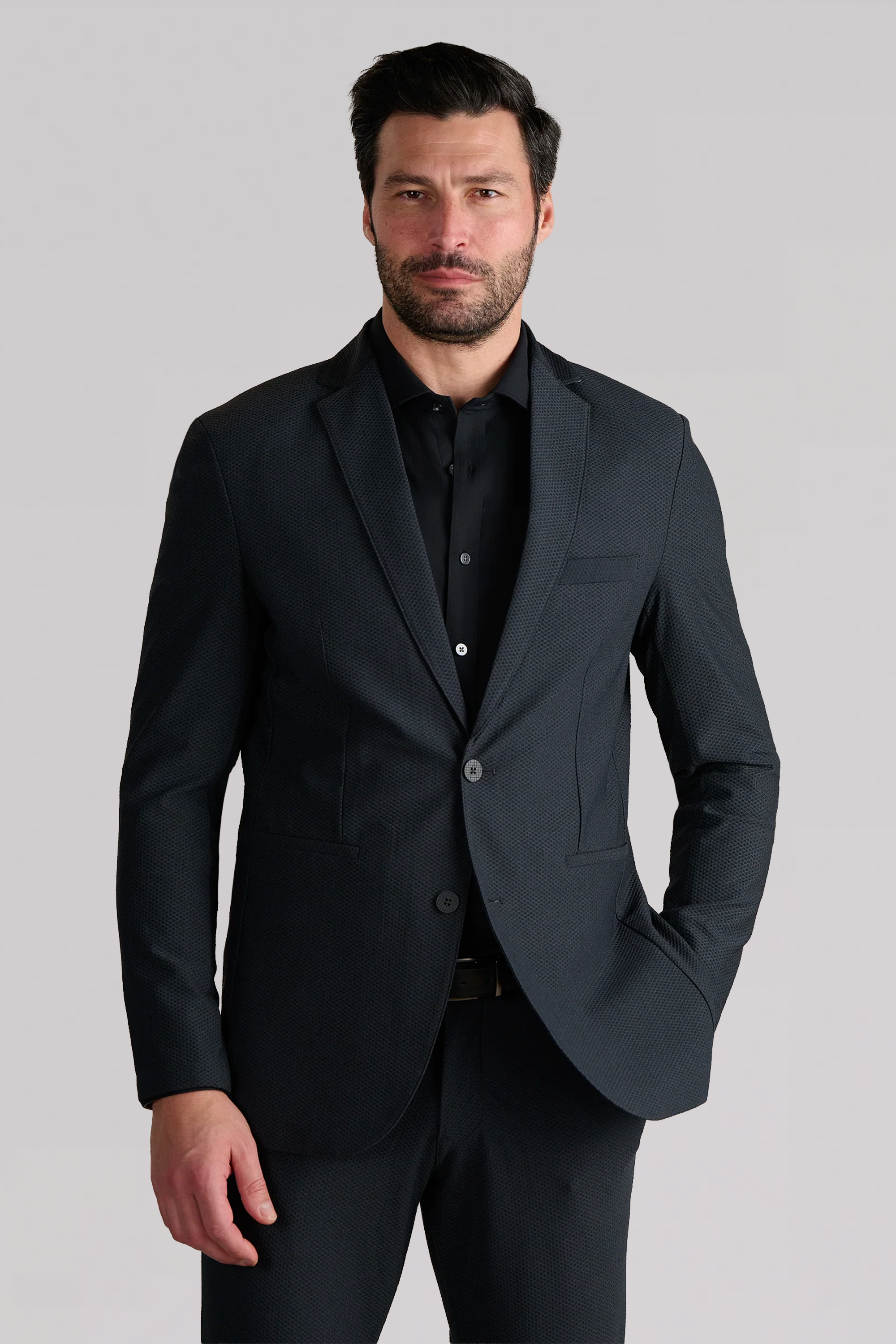 A man with short dark hair and a beard wears the Twillory AIR Blazer in Heather Charcoal over a black dress shirt. He faces forward with one hand in his pocket and a neutral expression, against a plain light gray background.