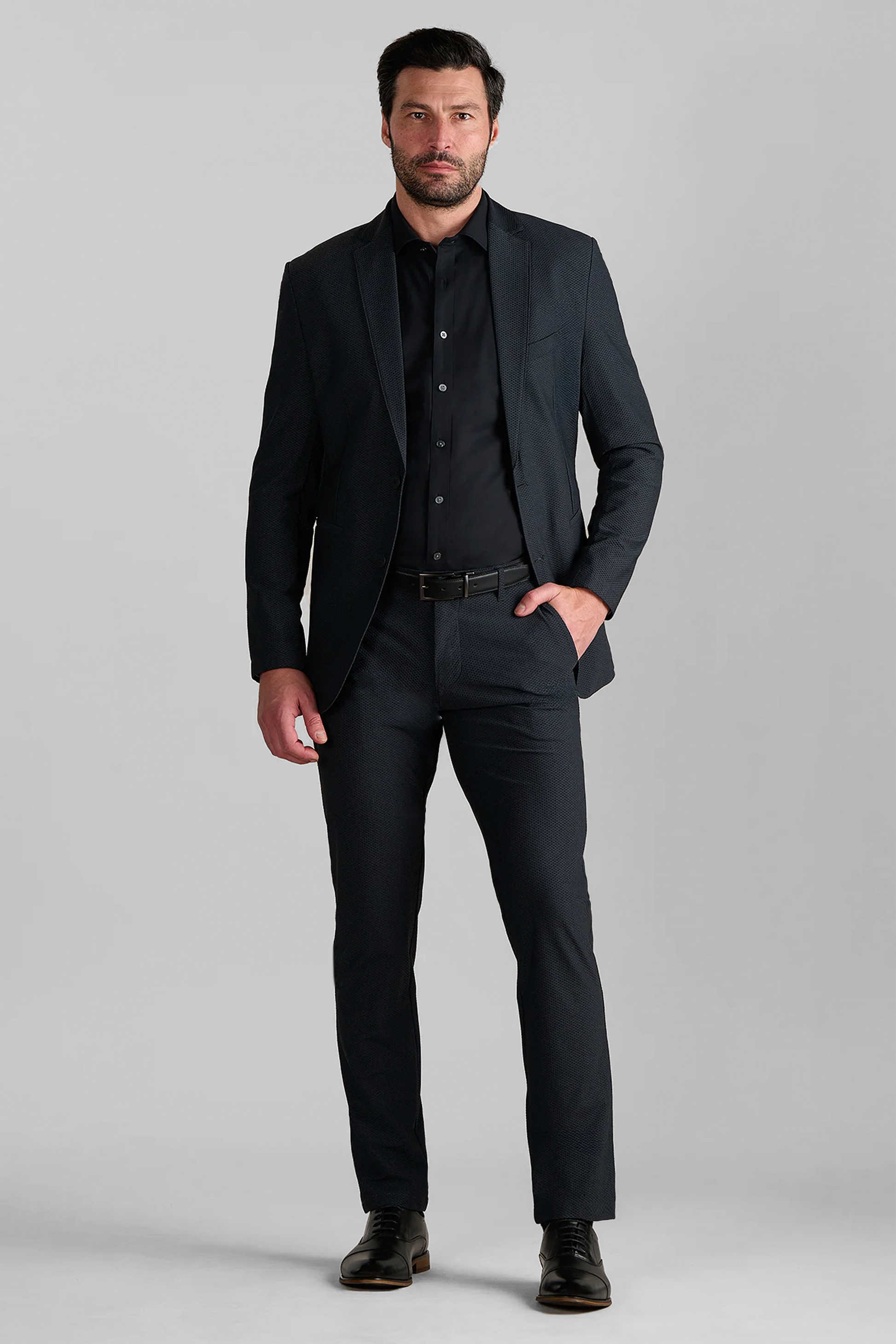 A man with short dark hair and a beard stands against a plain gray background, wearing Twillory's AIR Blazer in Heather Charcoal for a formal, professional look. One hand in his pocket, he pairs it with a black dress shirt and shoes.
