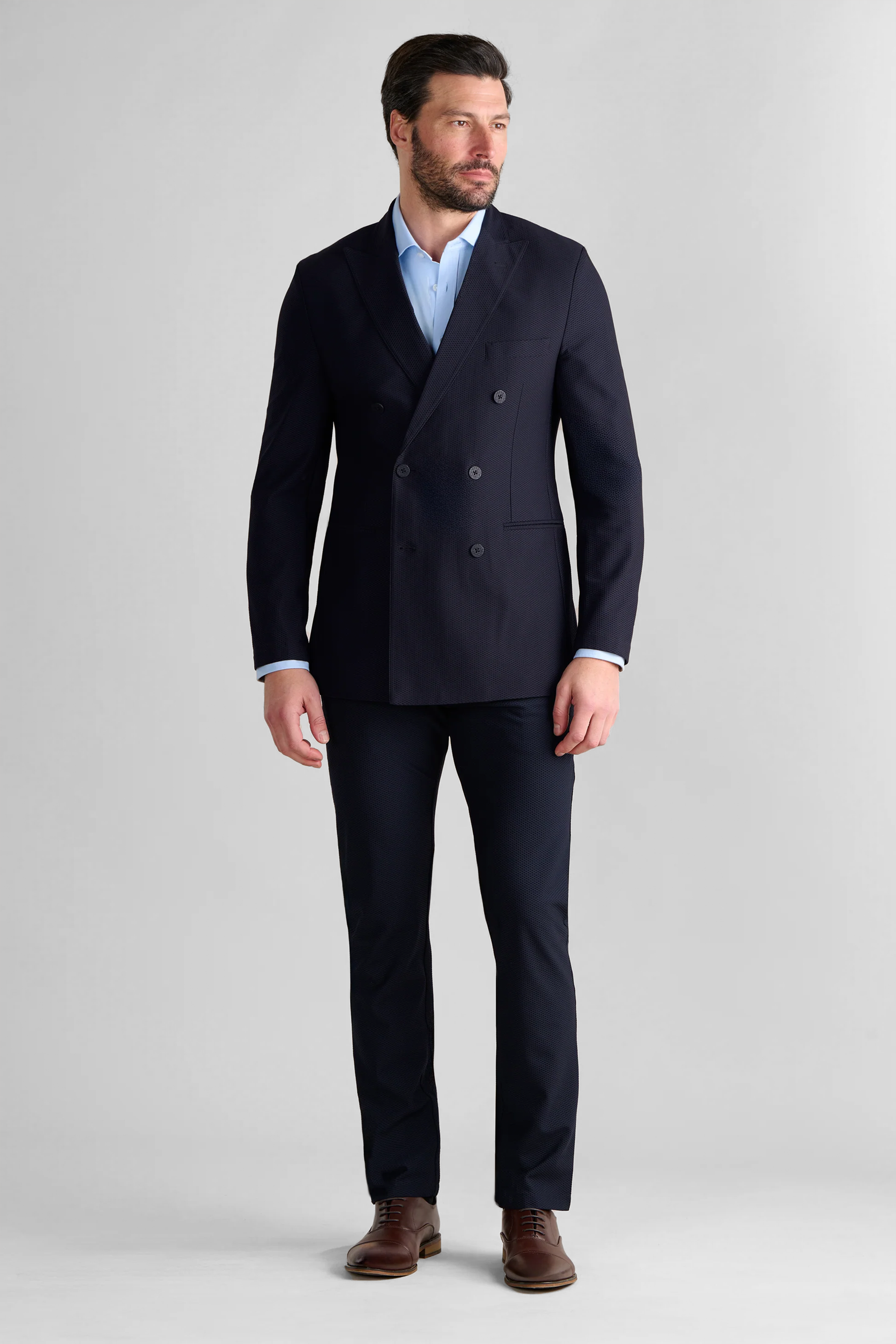 A man with short dark hair and a beard wears Twillory's AIR Double Breasted Blazer // Navy, made from 360° Airflow Fabric, over a light blue dress shirt with no tie and brown dress shoes, standing against a plain light gray background.