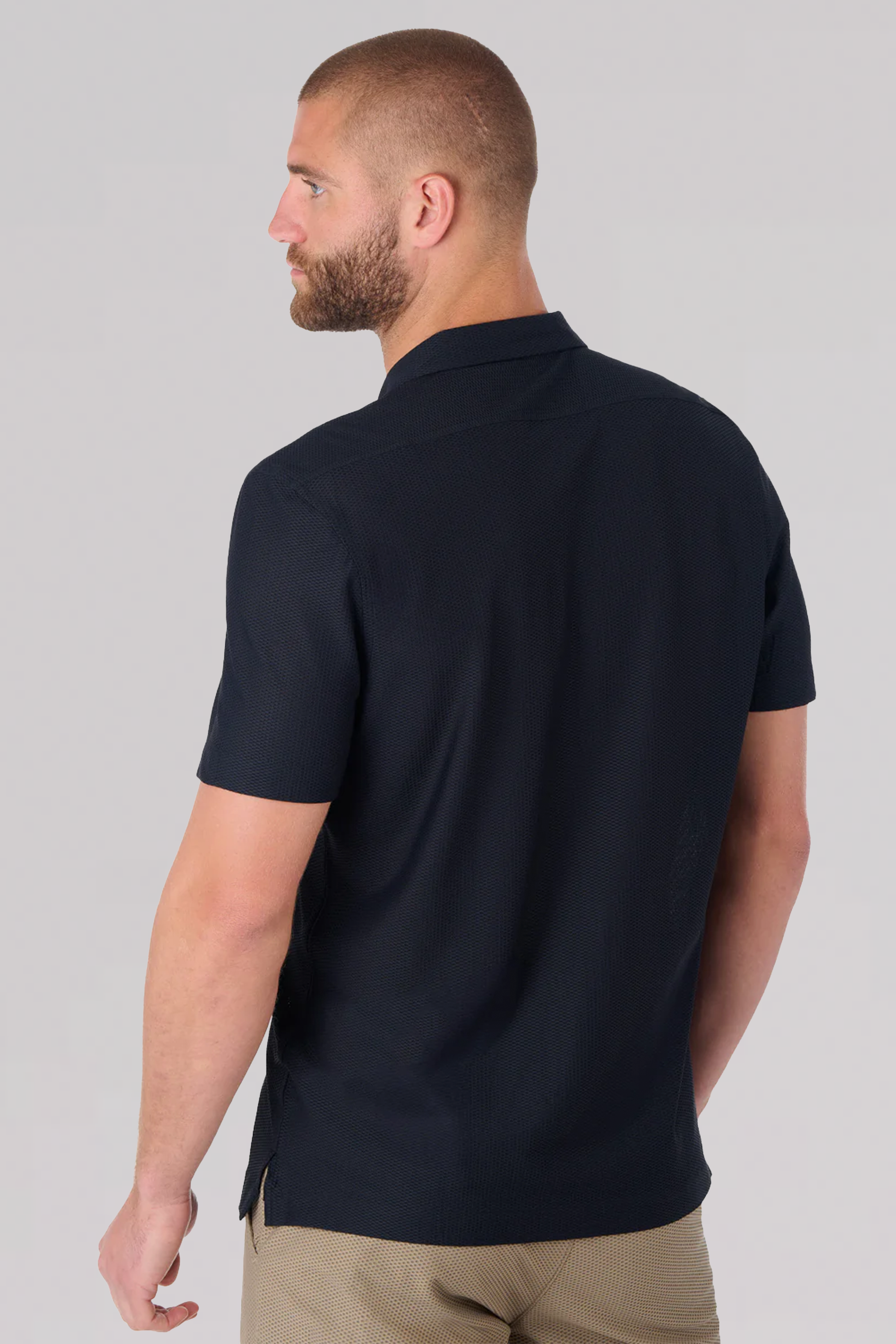 A man with short hair and a beard is seen from behind, wearing the Twillory AIR Resort Shirt Solid in Black with light brown pants, standing against a plain light gray background and turning his head slightly to the left.