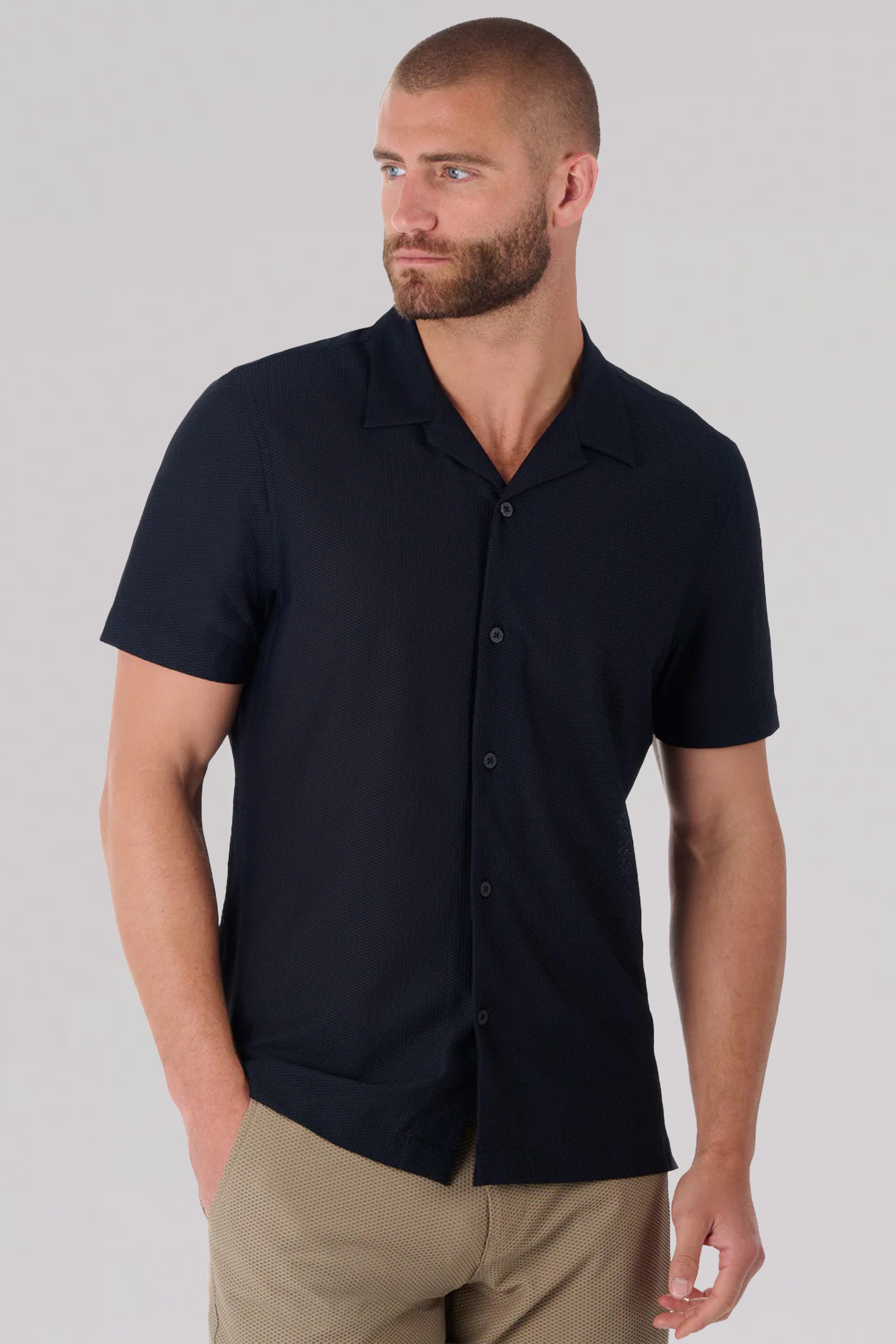 A man with short brown hair and a beard wears the Twillory AIR Resort Shirt Solid in black, paired with light brown pants. He faces slightly left with one hand in his pocket against a plain light gray background.
