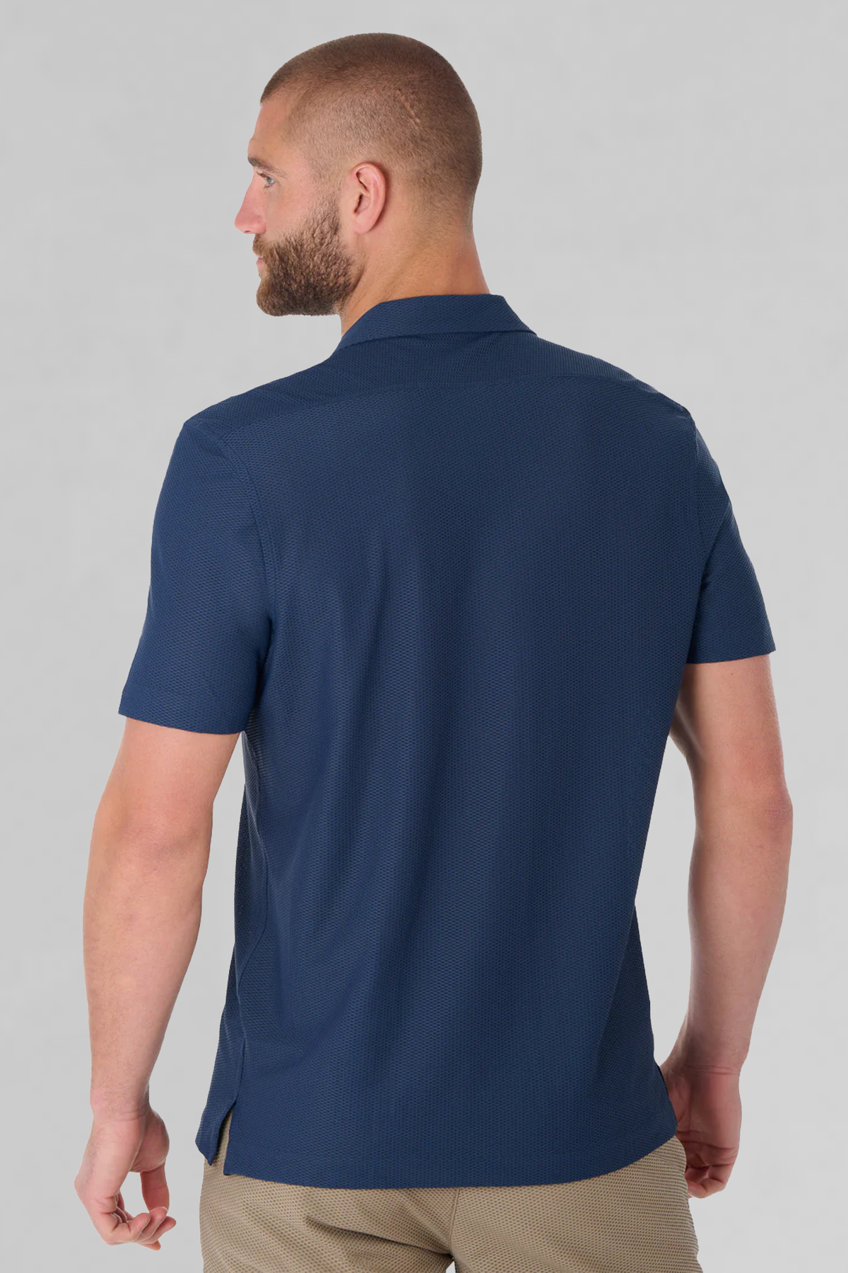 A man with short hair and a beard is shown from behind, wearing the Twillory AIR Resort Shirt Solid in navy with light brown pants. The shirt features a straight hem and side vents. The background is plain light gray.