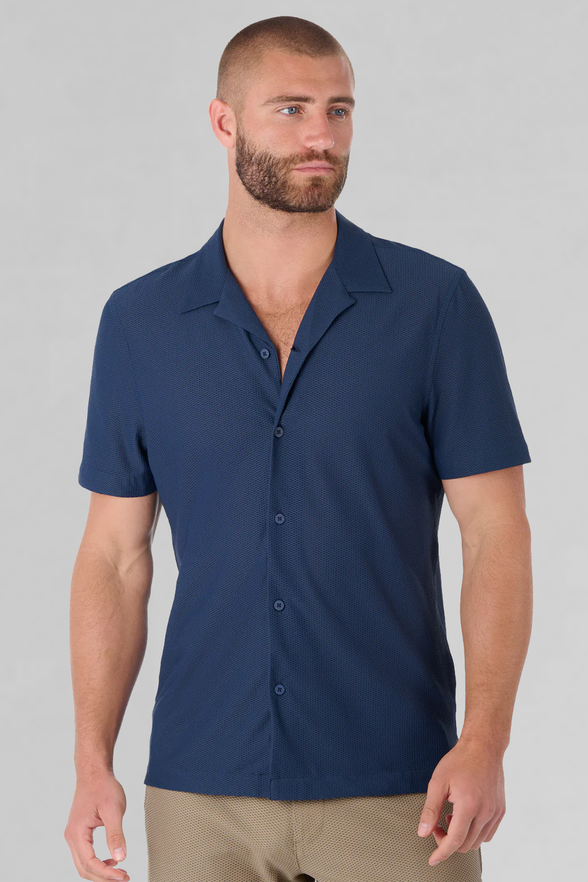 A man with short hair and a beard models the Twillory AIR Resort Shirt Solid in Navy, featuring short sleeves and cooling machine-washable fabric. He stands against a light gray background, looking to the side with a neutral expression. Only his upper body is visible.