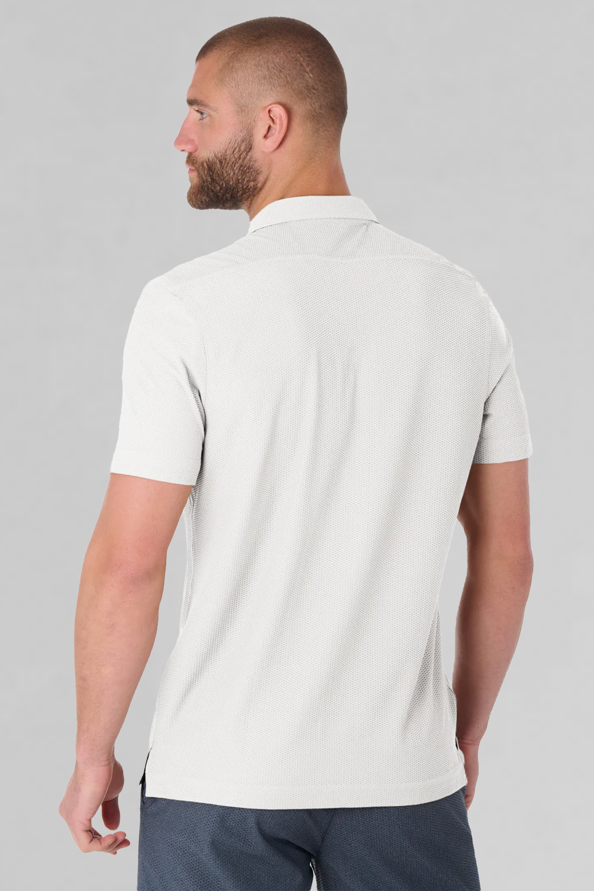 A man with a close-cropped beard and short hair, seen from behind, wears Twillory's AIR Resort Shirt Solid in white—crafted with cooling AIR fabric for breathability—paired with dark blue pants against a plain light gray backdrop.