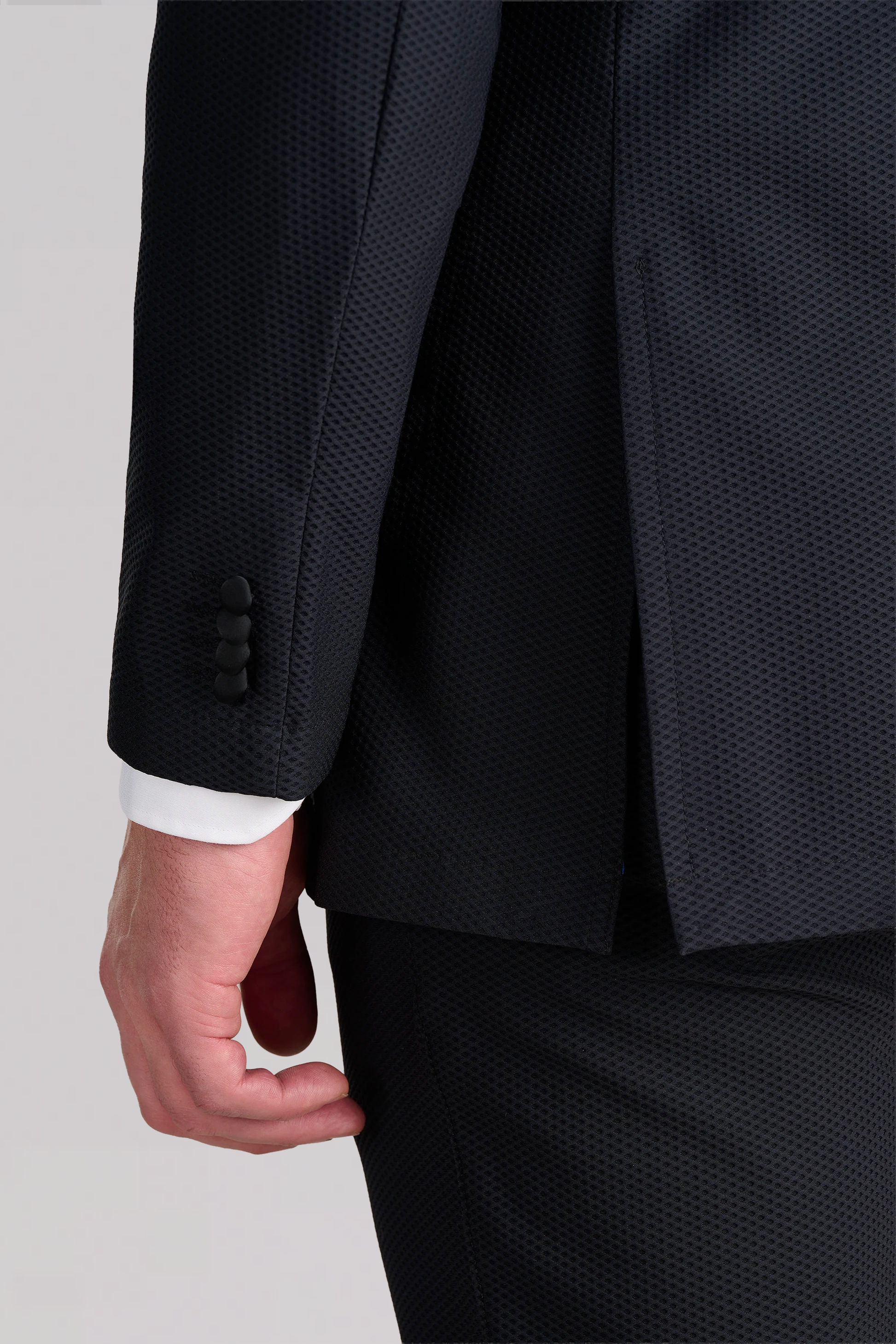 A close-up of a person wearing the Twillory AIR Tuxedo Blazer in black, featuring a textured fabric and visible white shirt cuff with three black buttons. Their arm hangs at their side against a light gray background.