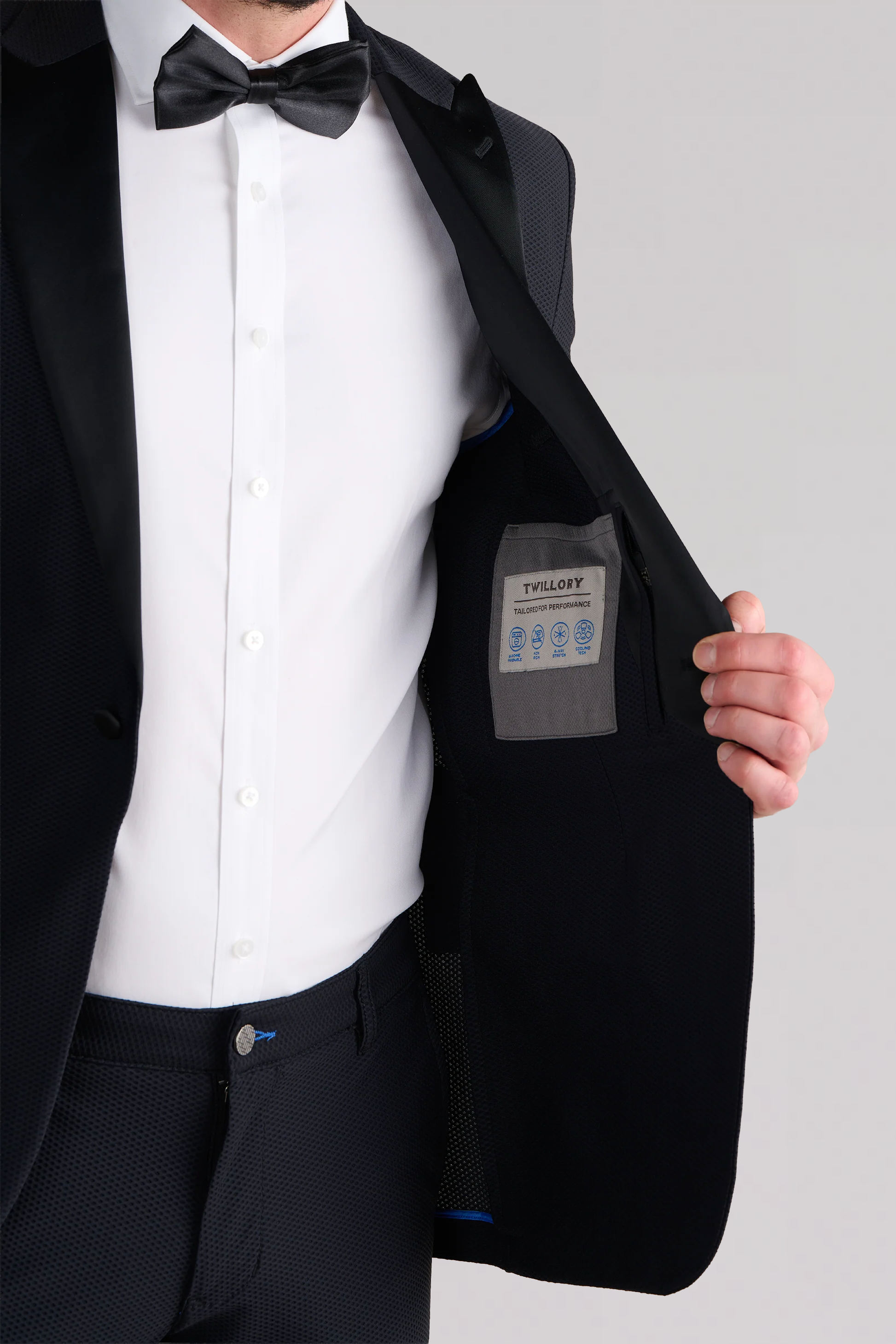 A man in a breathable Twillory AIR Tuxedo Blazer // Black holds his jacket open, revealing the gray inner pocket with a blue TWILORY label. The background is plain and light-colored.