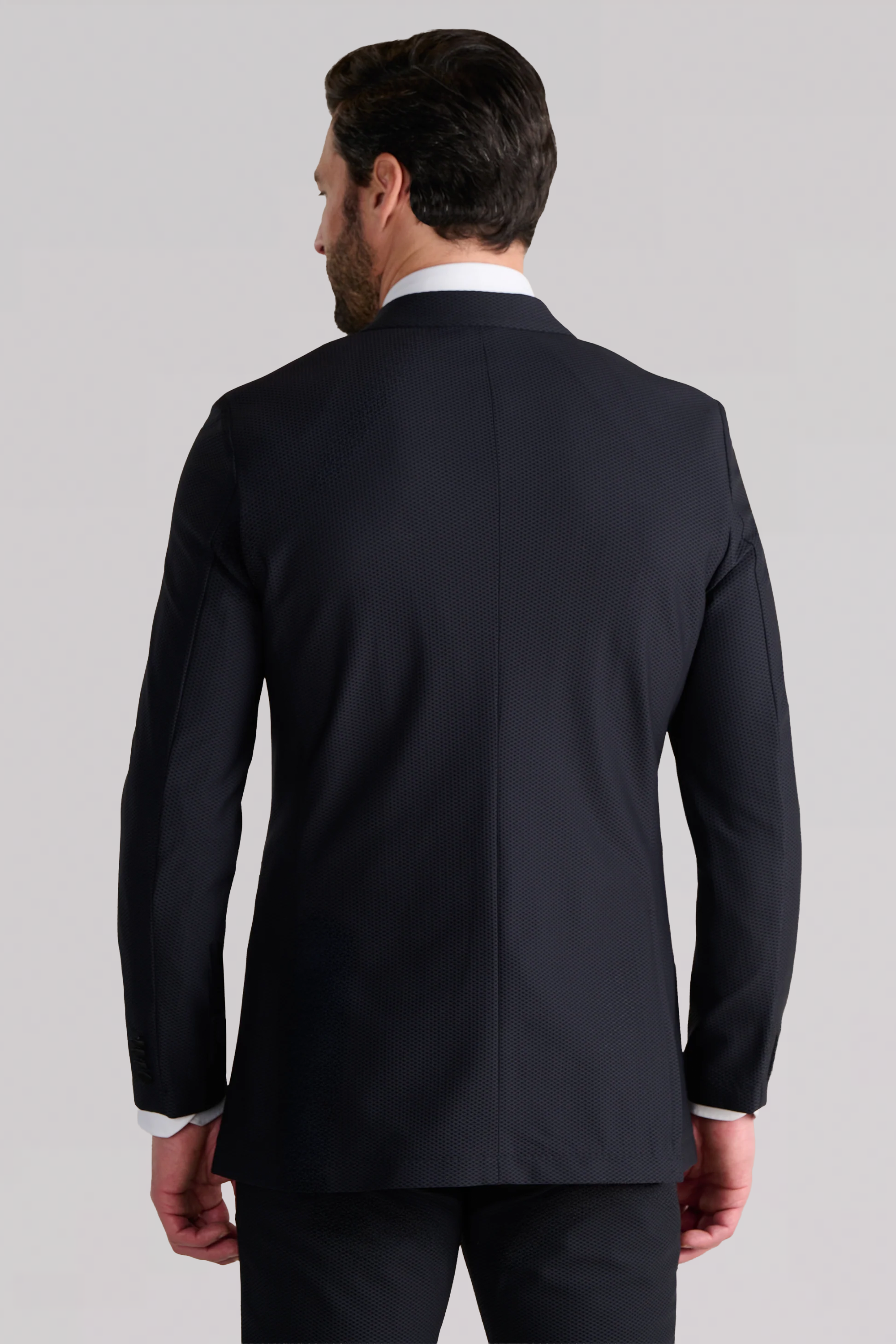A man with short brown hair is shown from behind, wearing the Twillory AIR Tuxedo Blazer in black with matching trousers and a white dress shirt. The plain, light background highlights this formal look.