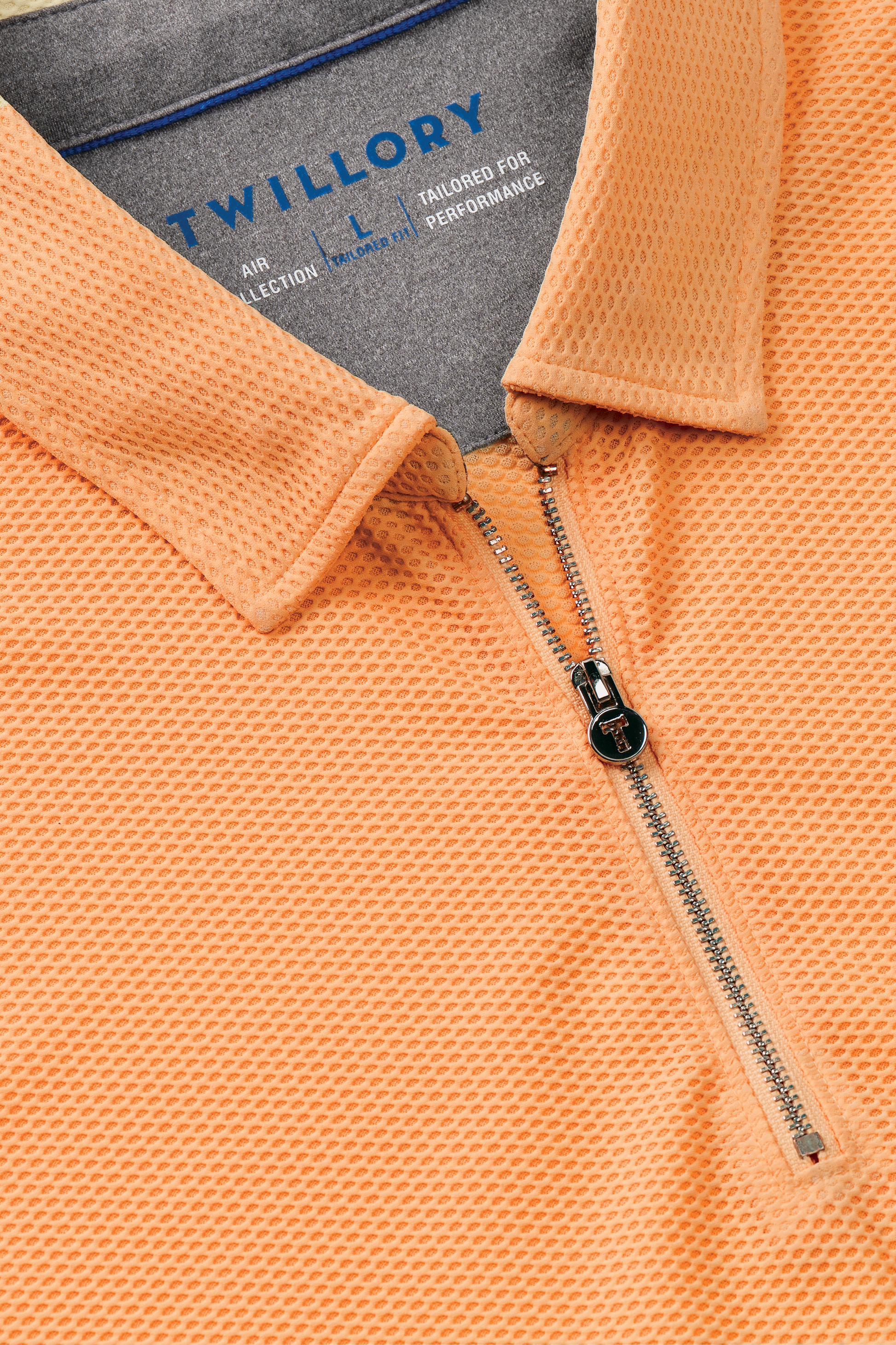 Close-up of the Twillory AIR Zip Polo Solid in orange, featuring a textured hyper-breathable fabric, silver quarter-zip with black pull tab, and an inside label detailing "Twillory AIR COLLECTION" and 360° Airflow Fabric.