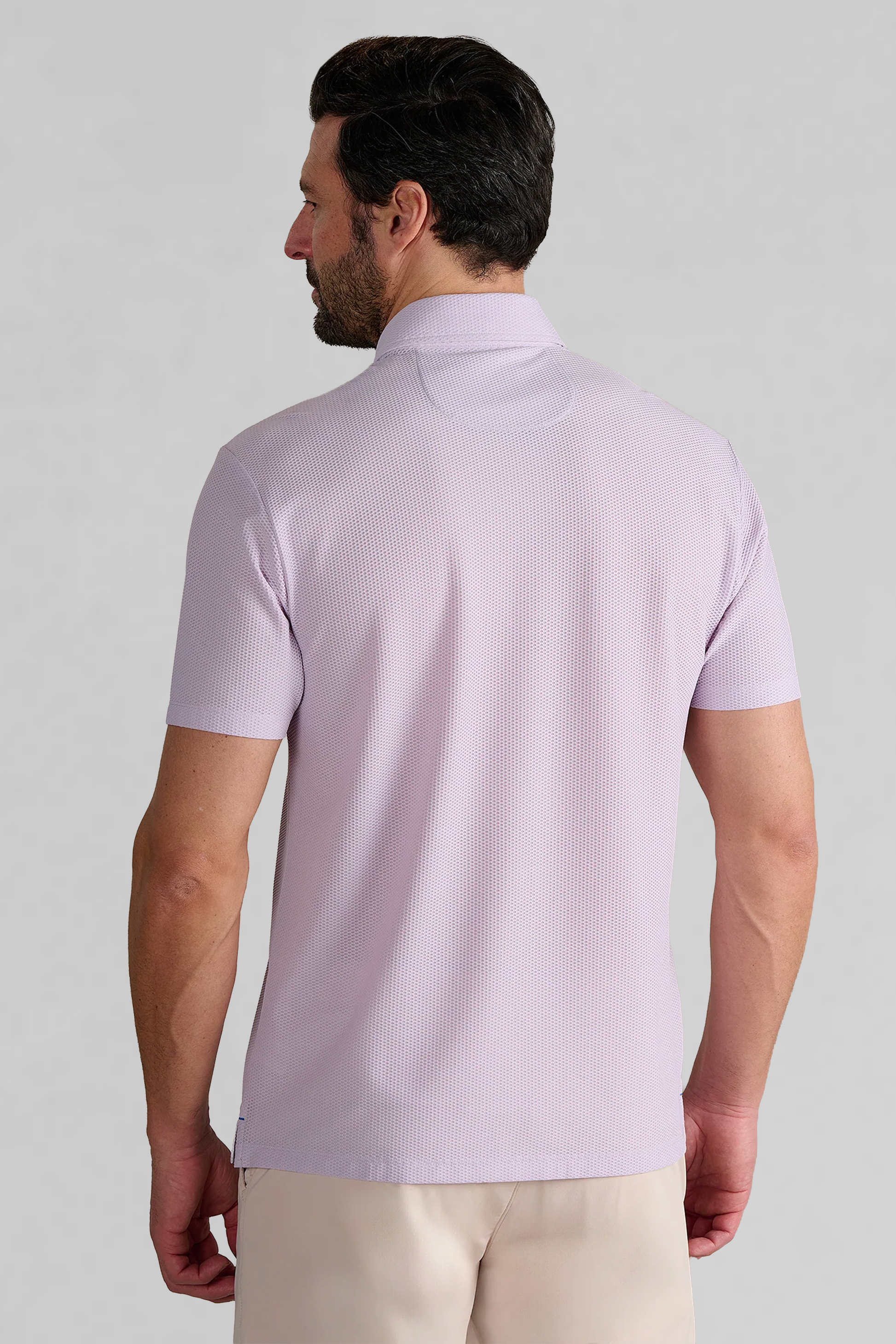 A man with dark, neatly styled hair and a trimmed beard faces away, wearing the Twillory AIR Zip Polo Solid in Lilac—a hyper-breathable, subtly textured polo with a classic collar—against a plain light gray background.