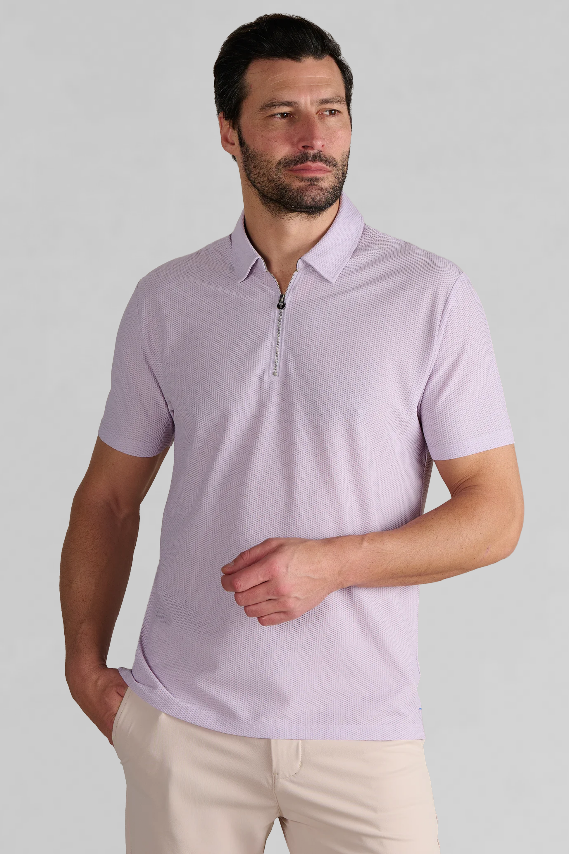 A man with short dark hair and a beard wears the Twillory AIR Zip Polo Solid in Lilac and beige pants. He stands against a plain light gray background, looking slightly to the side with one hand in his pocket.