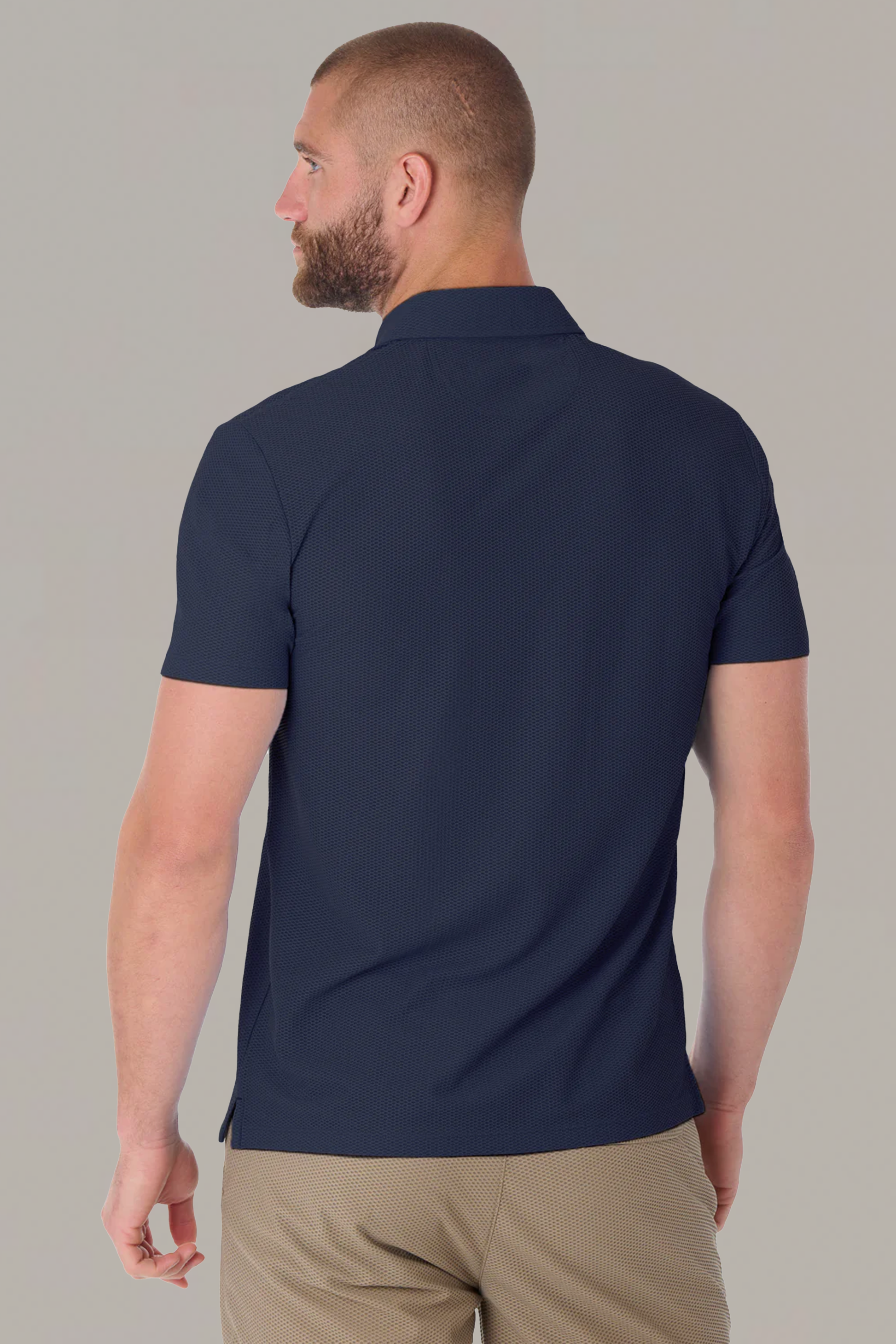 A man with short hair and a beard is seen from behind wearing the Twillory AIR Zip Polo Solid in Navy and light brown pants. The polo features 360° Airflow Fabric for hyper-breathability. He stands before a plain, light gray background.