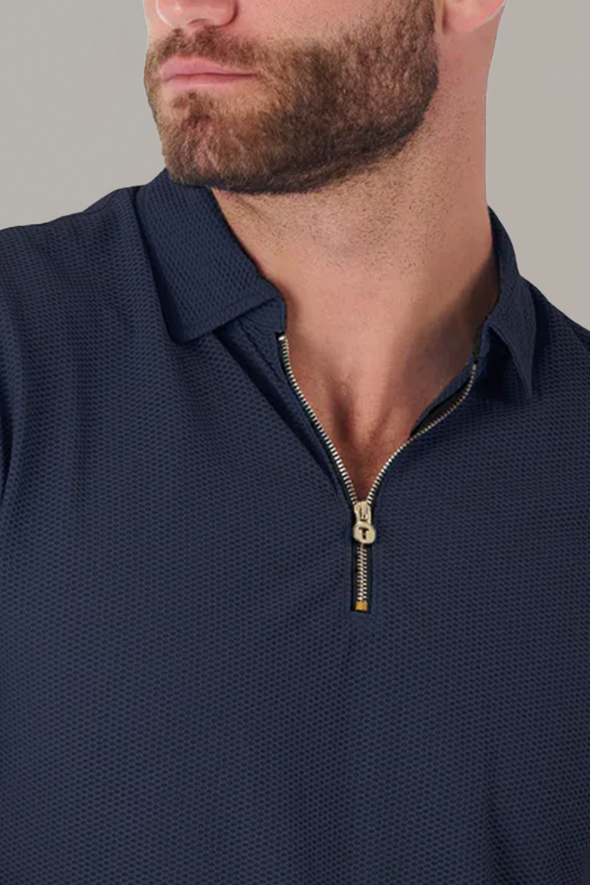 A close-up of a man’s lower face and upper torso shows him wearing the Twillory AIR Zip Polo Solid in navy with a textured finish and silver zipper, set against a plain light grey background. He has short facial hair.