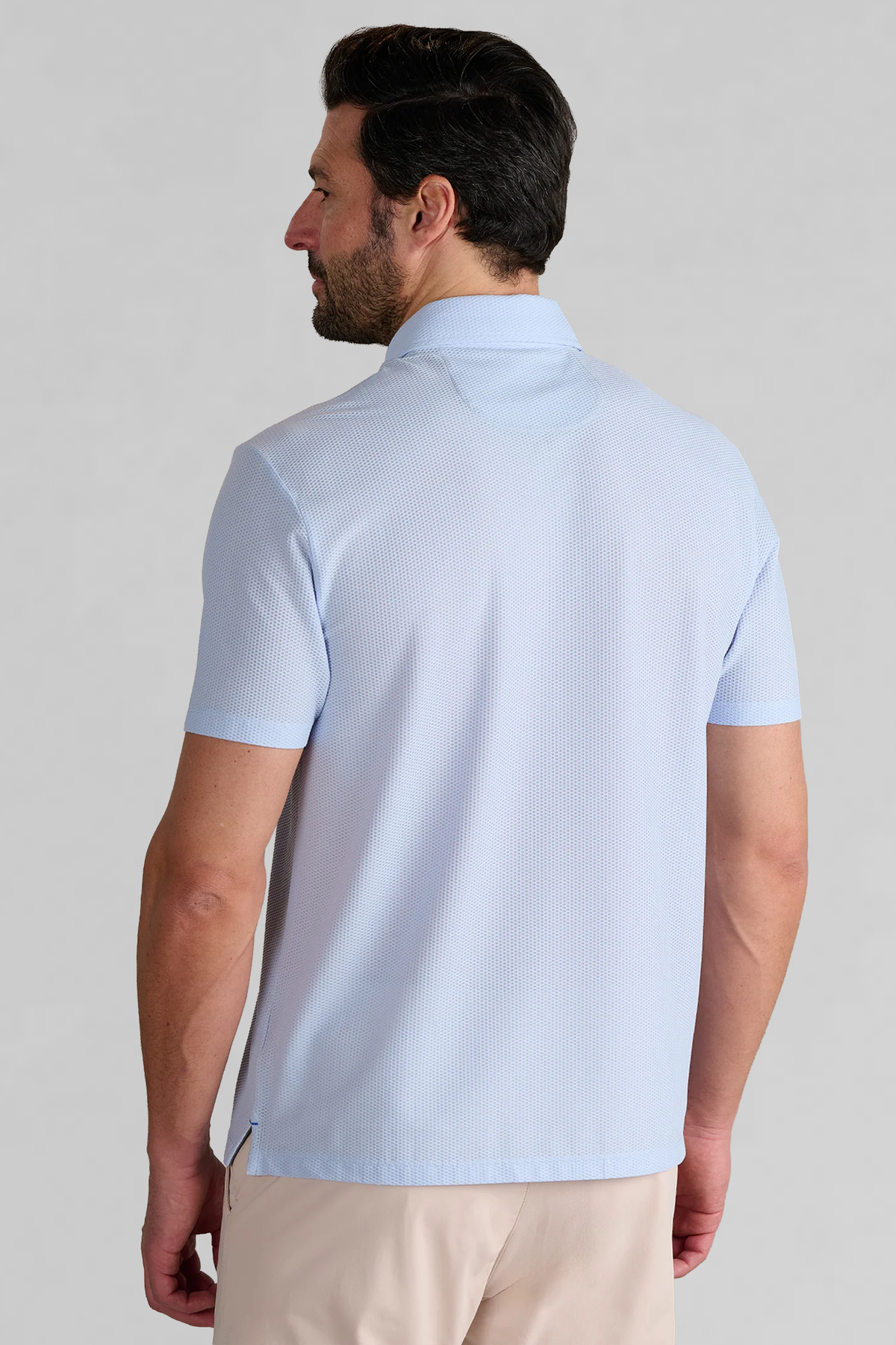 A man faces away from the camera in a Twillory AIR Zip Polo Solid in Powder Blue—a hyper-breathable polo with 360° Airflow Fabric, textured collar, and beige pants—his dark hair styled neatly against a plain light gray background.