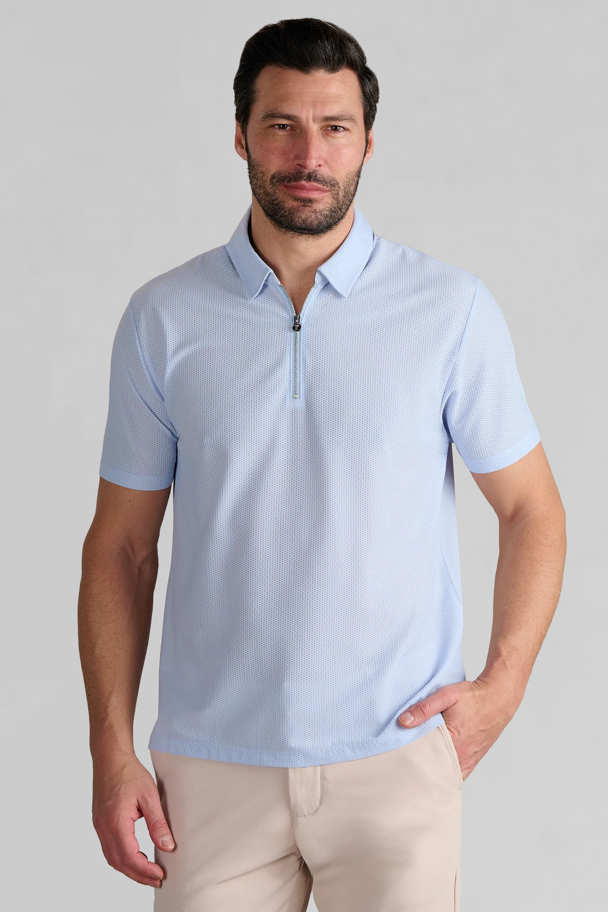 A man with short dark hair and a beard wears the Twillory AIR Zip Polo Solid in Powder Blue, featuring short sleeves and a zip-up collar. He stands against a plain gray background with one hand in his pocket and a neutral expression.