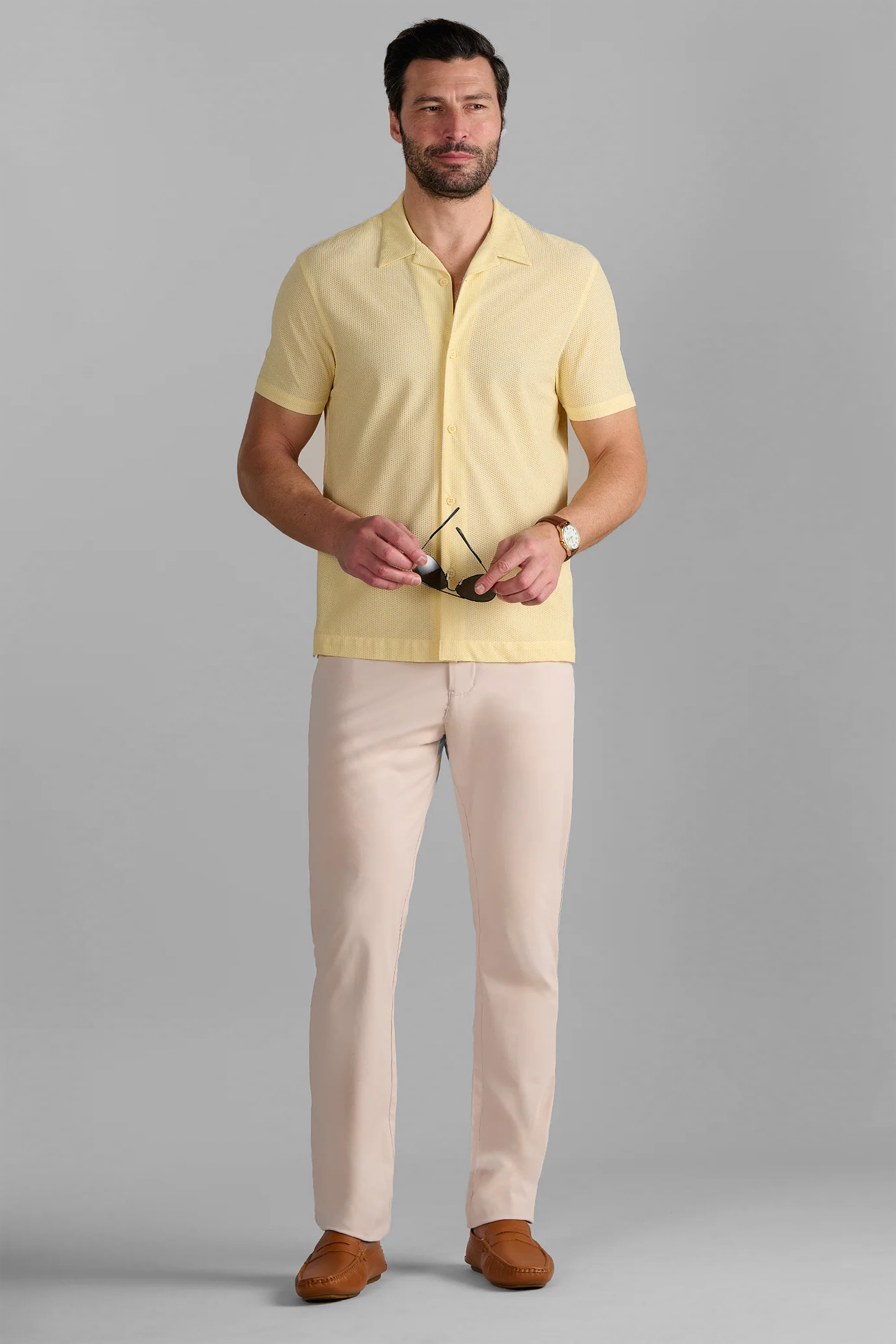 A man with short dark hair, a beard, and a slight smile stands against a gray background wearing the Twillory AIR Resort Shirt Solid in Yellow, pale beige pants, and brown loafers. He holds sunglasses and wears a wristwatch.