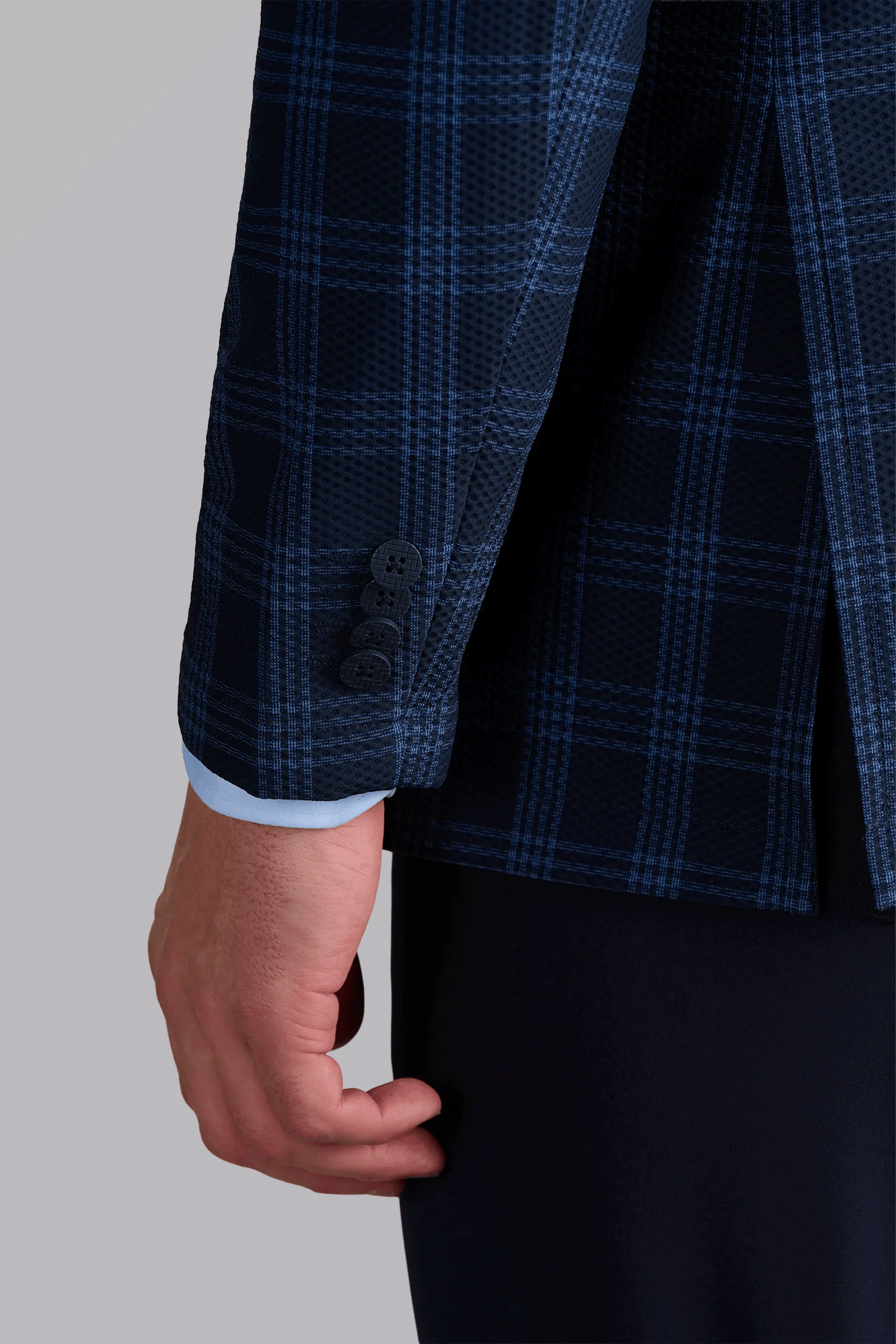 A close-up of a person’s left arm in the Twillory AIR Blazer // Navy Blue Plaid, featuring a dark blue plaid, machine-washable finish, subtle blue check pattern, black buttons, and light blue shirt cuff against a gray background.