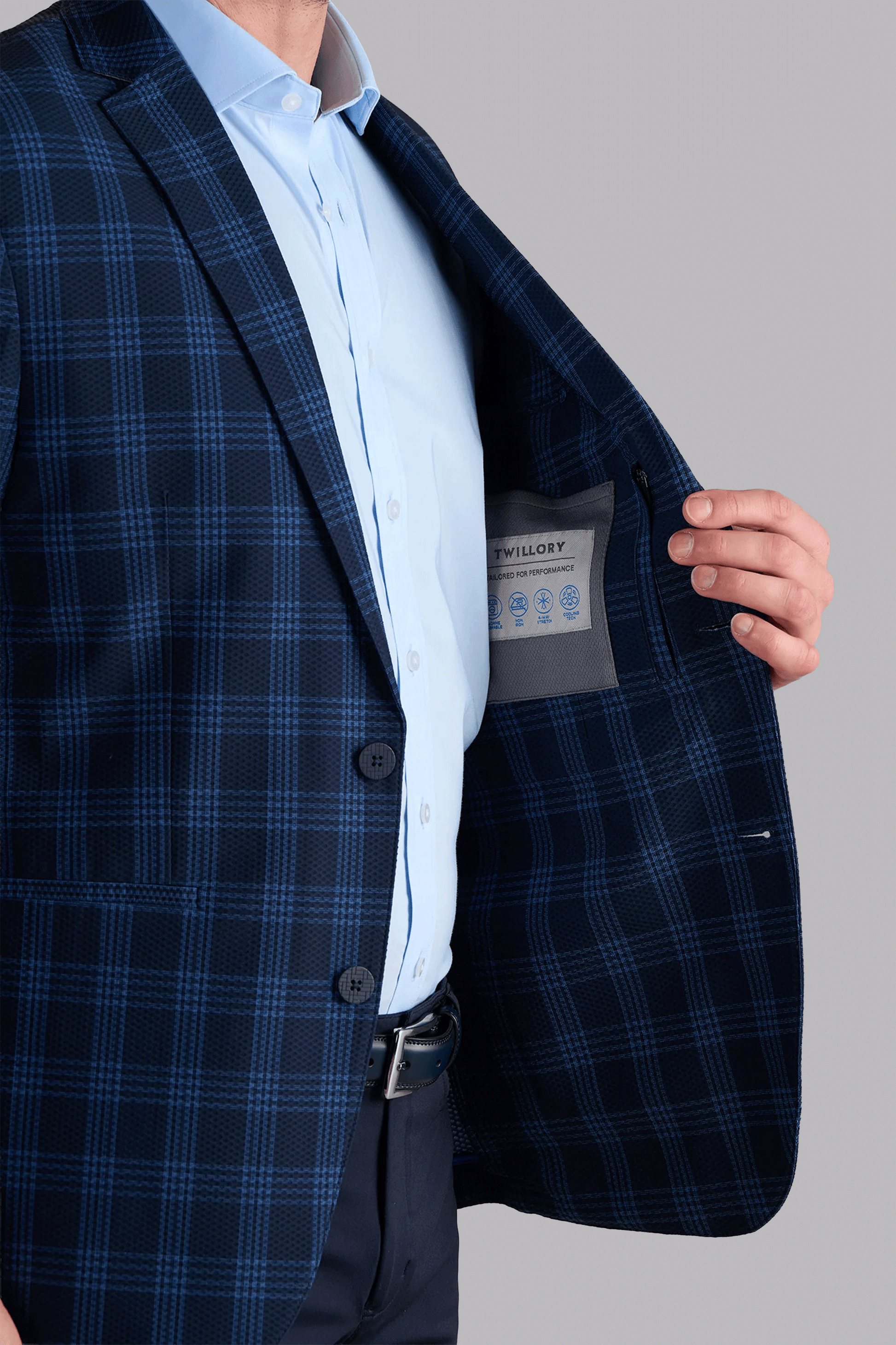 A man in a Twillory AIR Blazer // Navy Blue Plaid made with 360° Airflow Fabric and a light blue shirt opens the jacket to reveal its inner label, against a plain gray background. Only his torso, right arm, and hand are visible.