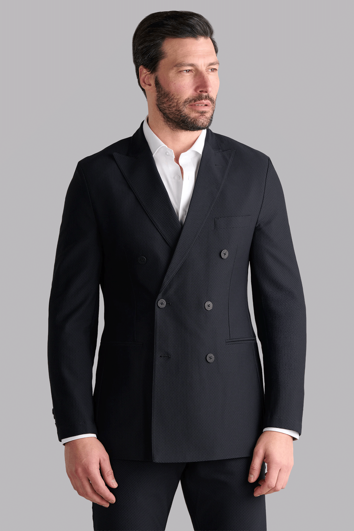 A man with short dark hair and a beard wears the Twillory AIR Double Breasted Blazer // Black over a white dress shirt. He stands against a plain light gray background, facing slightly right with his hands relaxed at his sides.