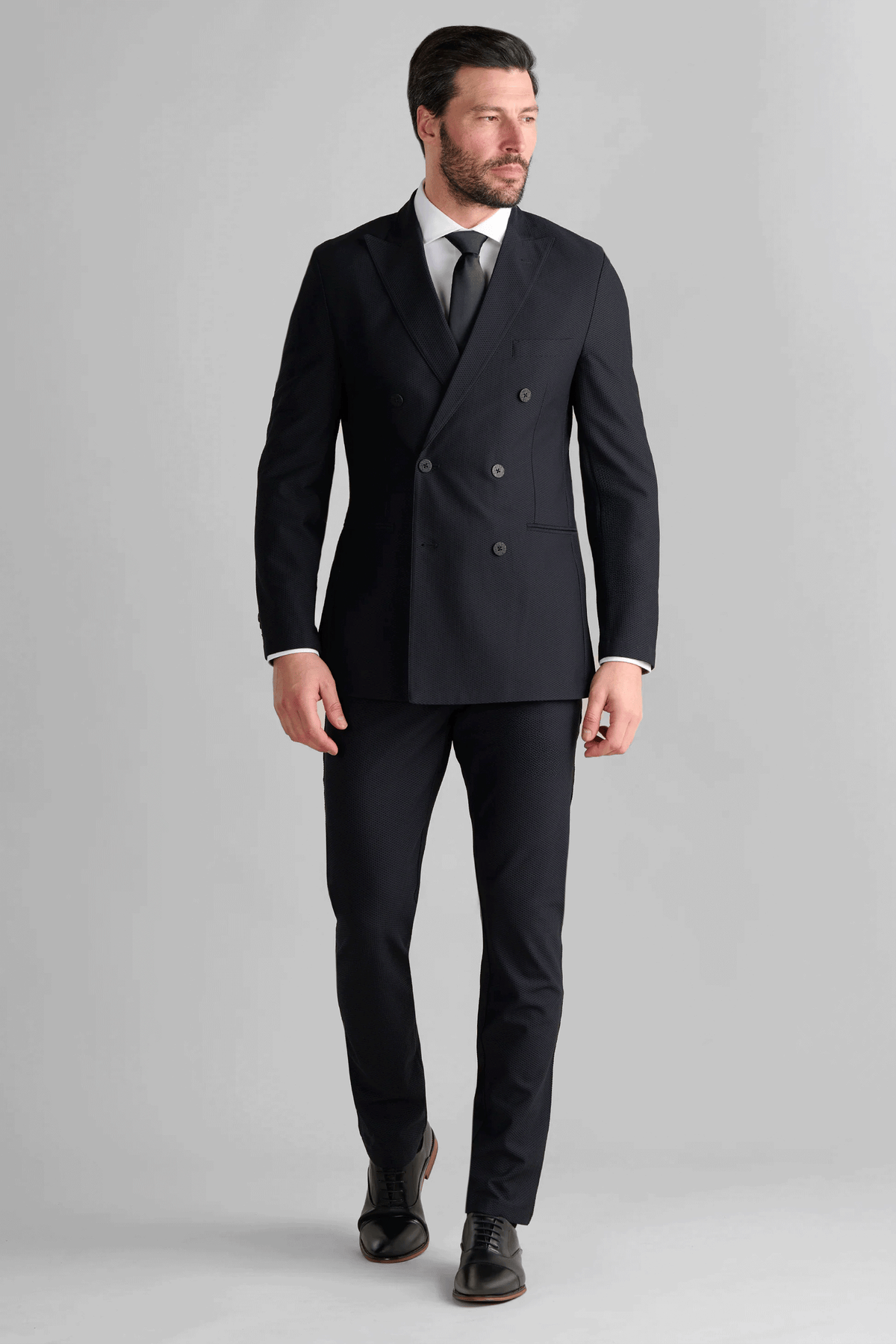 A man with short dark hair and a beard wears the Twillory AIR Double Breasted Blazer // Black, made from 360° Airflow Fabric, over a white dress shirt and black tie. He stands against a light gray background, looking slightly left, mid-stride.