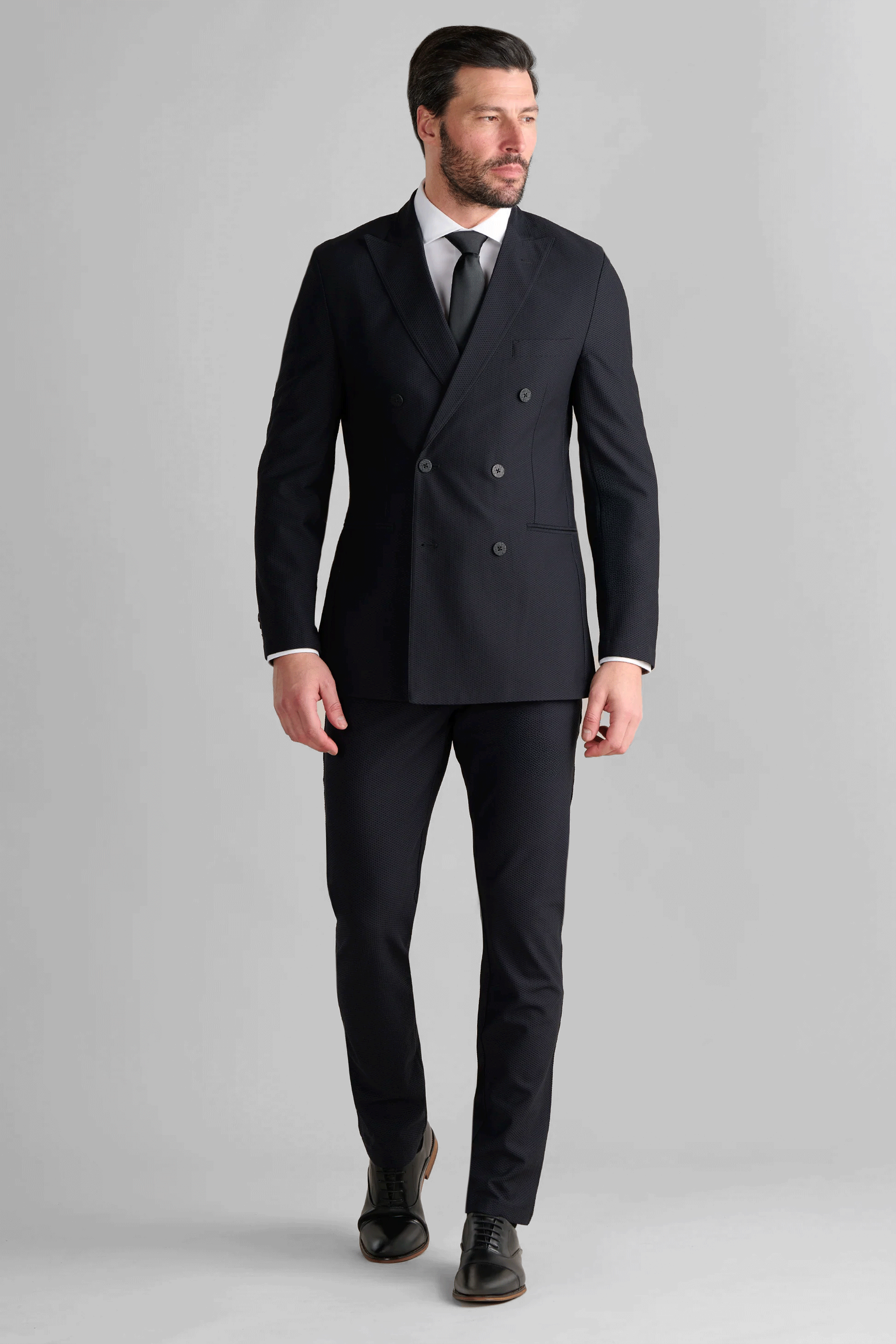A man with short dark hair and a beard wears the Twillory AIR Double Breasted Blazer // Black, made from 360° Airflow Fabric, over a white dress shirt and black tie. He stands against a light gray background, looking slightly left, mid-stride.