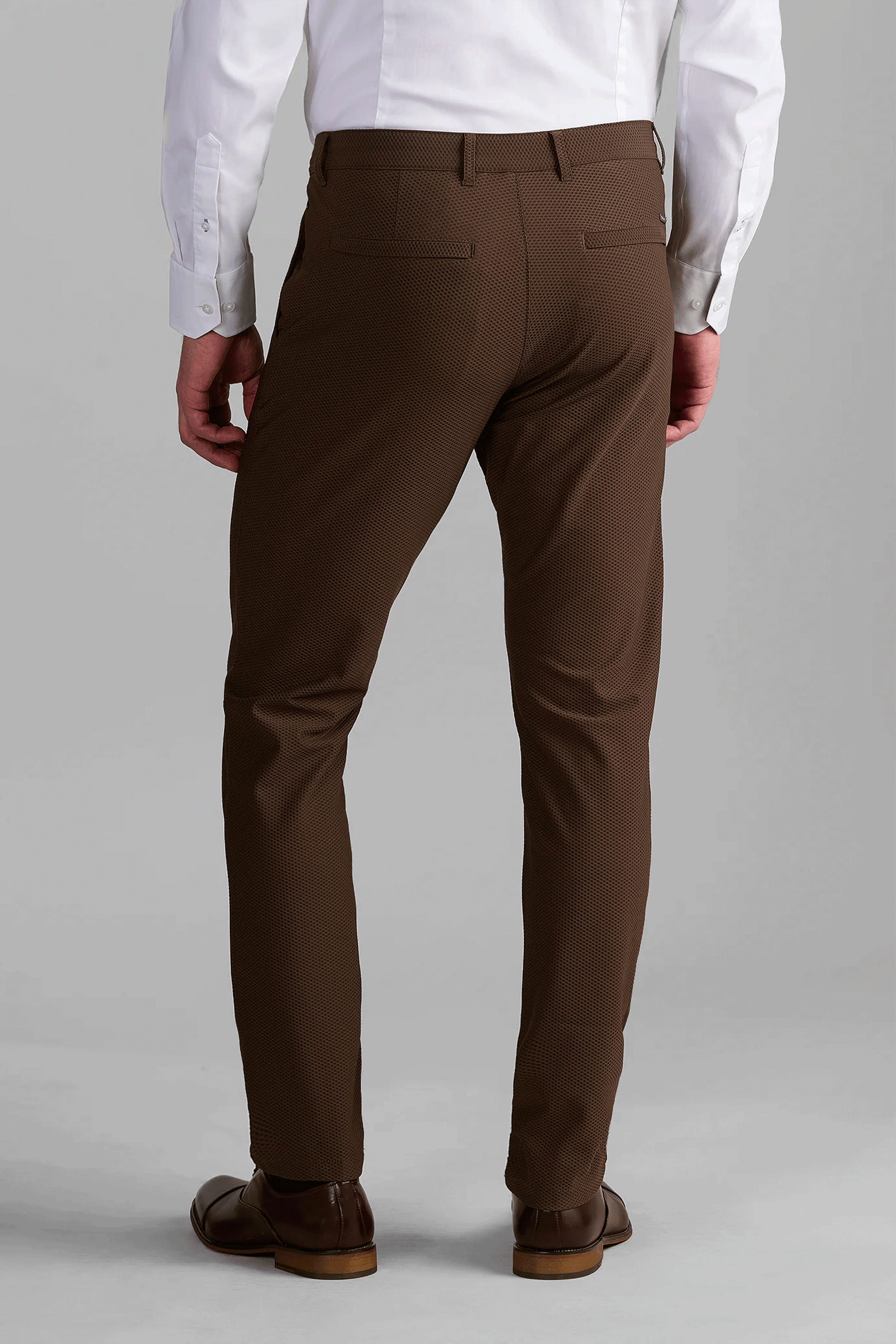 A man stands facing away in Twillory AIR Pants // Brown—slim-fit, breathable dress pants with two back welt pockets—paired with light brown dress shoes and a tucked-in white shirt. The background is plain light gray.
