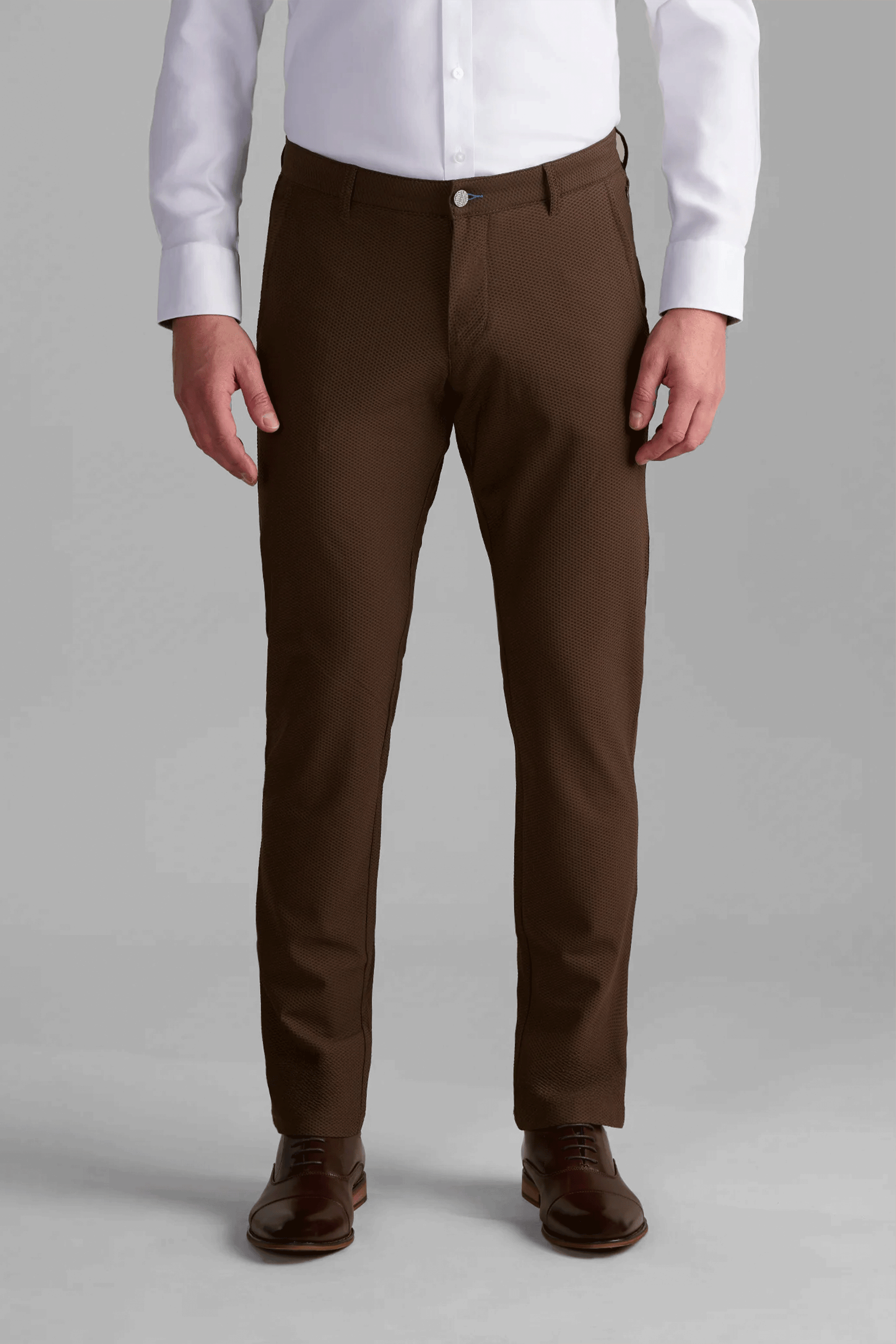 A person stands facing forward, wearing a white dress shirt tucked into Twillory AIR Pants // Brown, paired with dark brown leather dress shoes. Their hands are relaxed by their sides against a plain light gray background.