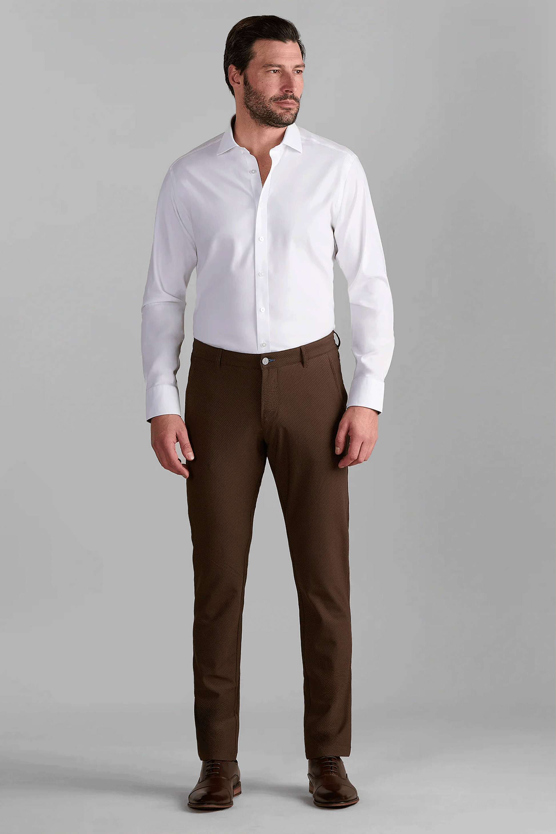 A man with short dark hair and a beard stands against a gray background, wearing Twillory AIR Pants in brown with a white dress shirt, belt, and brown dress shoes. He looks slightly to his right.