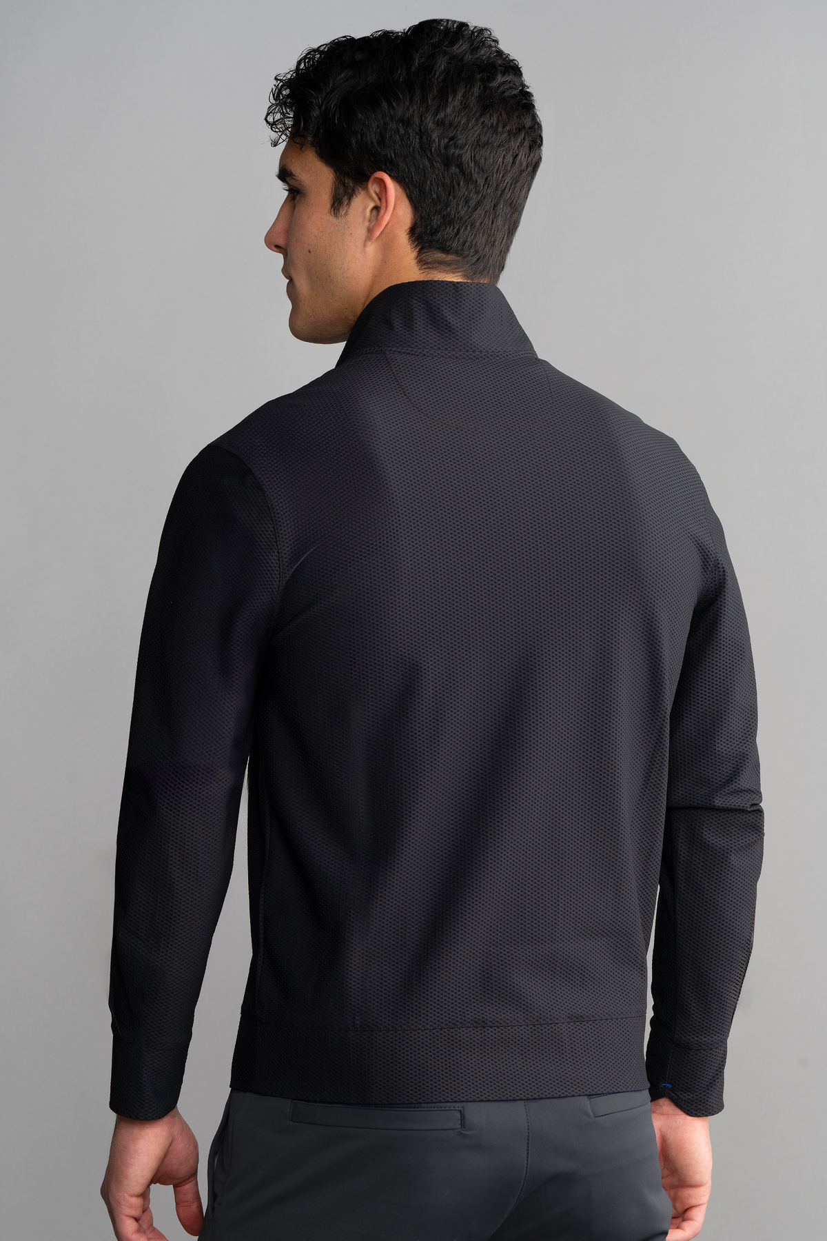A man with short, dark curly hair is seen from the back wearing the Twillory Air Quarter Zip in black, made of wrinkle-free stretch fabric. He stands against a plain gray background, looking slightly left with his left hand by his side.
