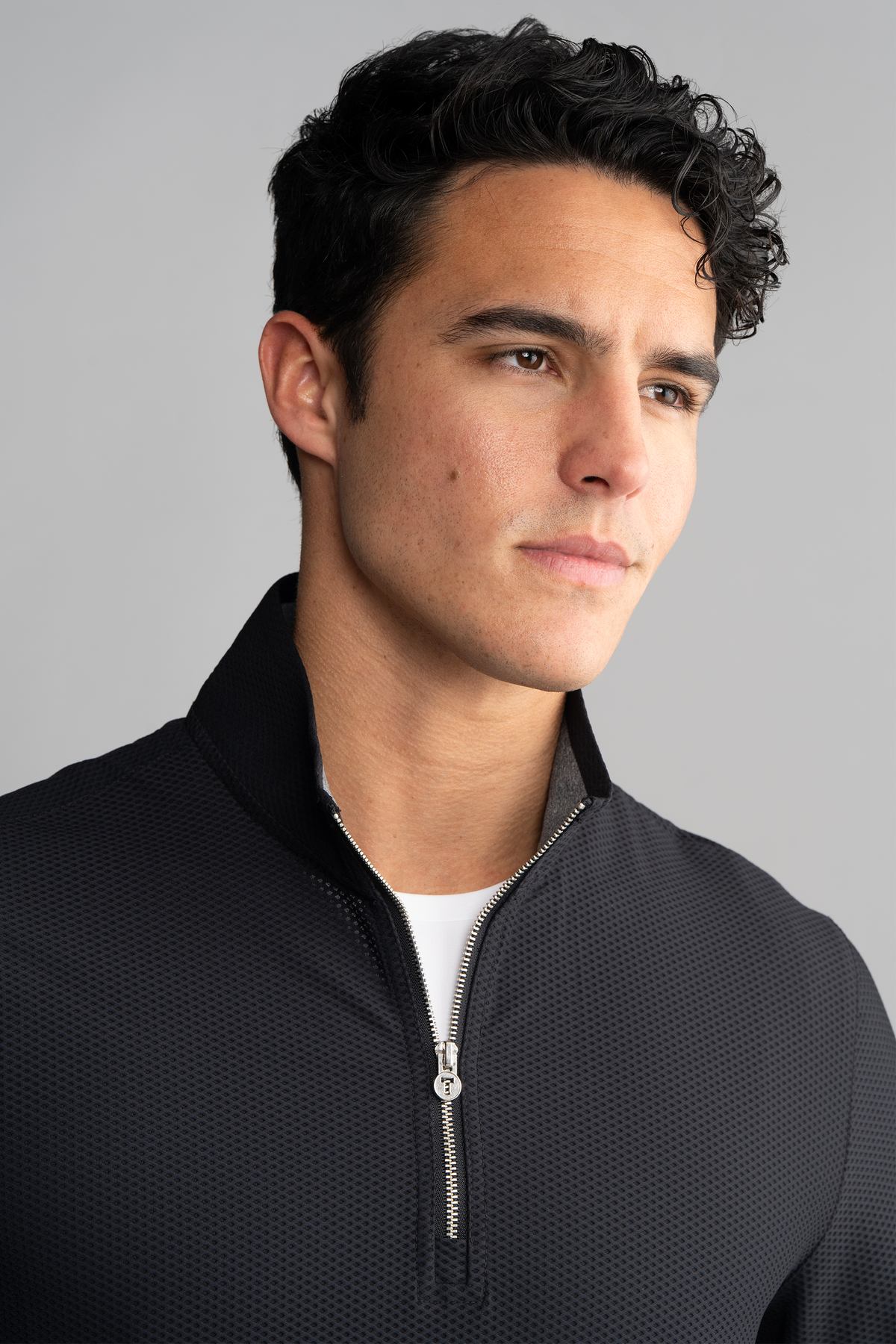A young man with short, dark curly hair and light skin looks thoughtfully to the side, wearing the Twillory Air Quarter Zip in black over a white shirt. The background is plain gray, and his expression is calm and composed.