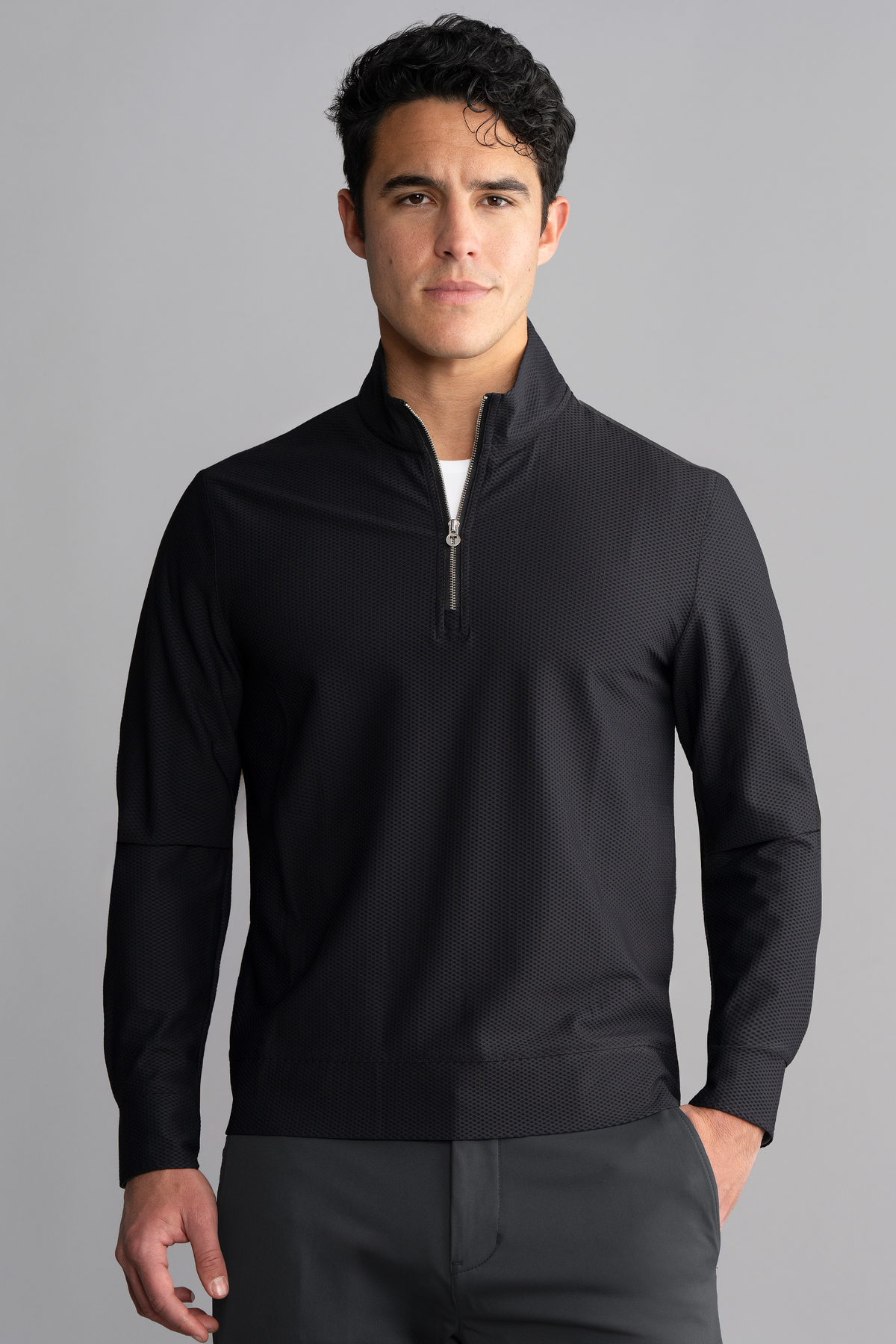 A man with short, wavy dark hair wears the Twillory Air Quarter Zip in black and dark pants. He stands facing forward against a plain gray background, hands at his sides, looking directly at the camera with a neutral expression.