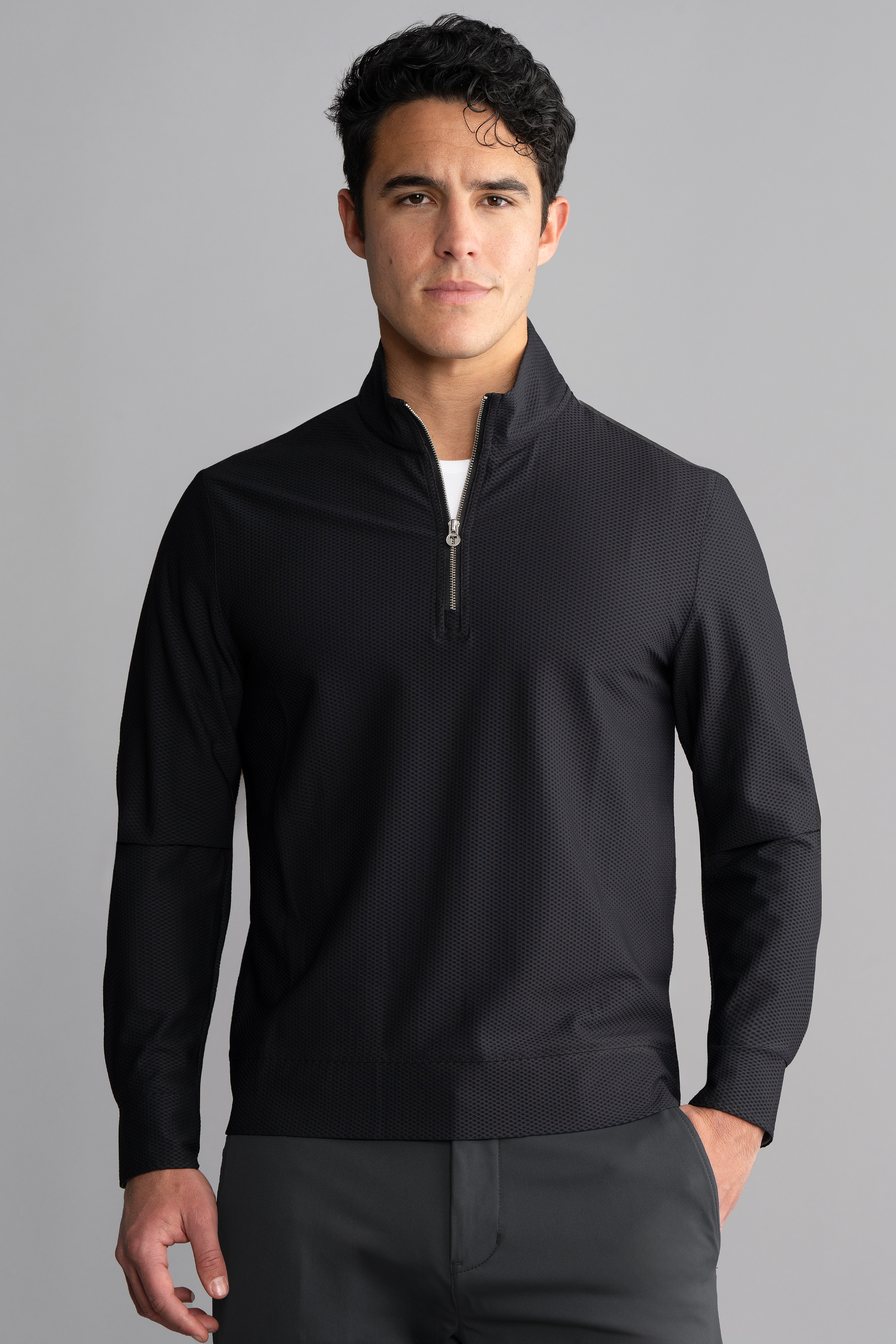 A man with short, wavy dark hair wears the Twillory Air Quarter Zip in black and dark pants. He stands facing forward against a plain gray background, hands at his sides, looking directly at the camera with a neutral expression.