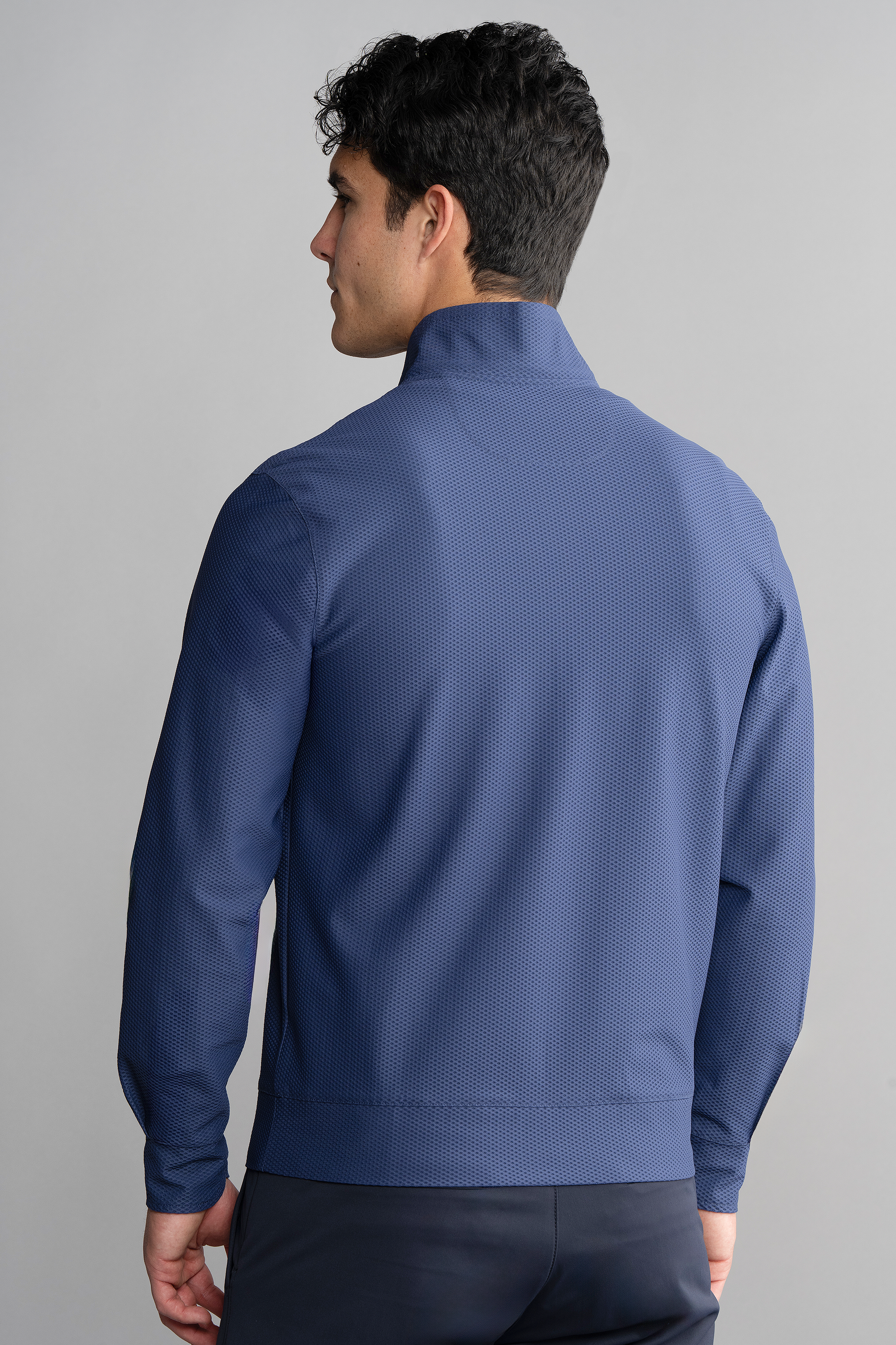 A man with short, dark, curly hair is seen from behind wearing the Twillory Air Quarter Zip in blue. The fitted, wrinkle-free stretch jacket stands out against a plain light gray background, emphasizing its smooth texture and vibrant color.