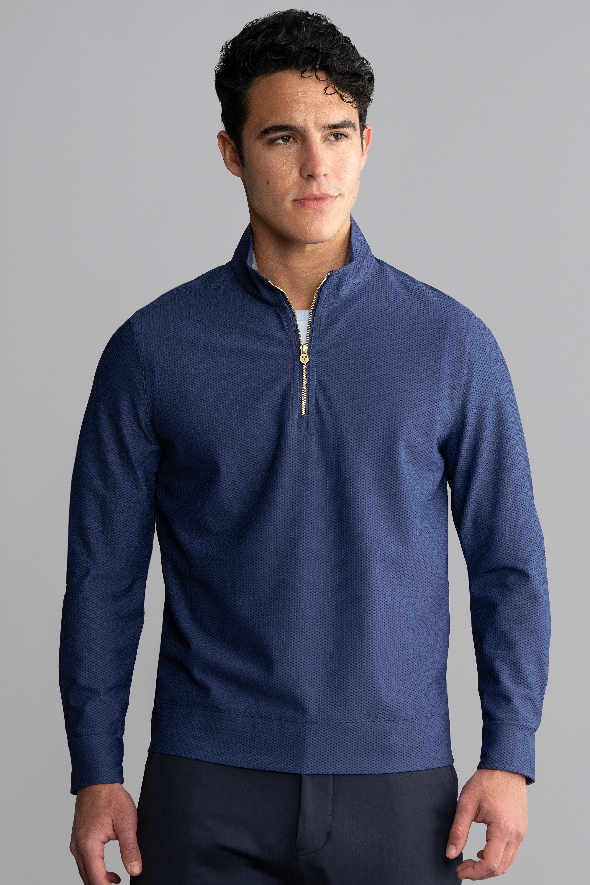 A young man with short, dark, wavy hair wears Twillory's Air Quarter Zip in blue, made from wrinkle-free stretch fabric. He stands facing forward against a plain light gray background, looking slightly to the left with a neutral expression.
