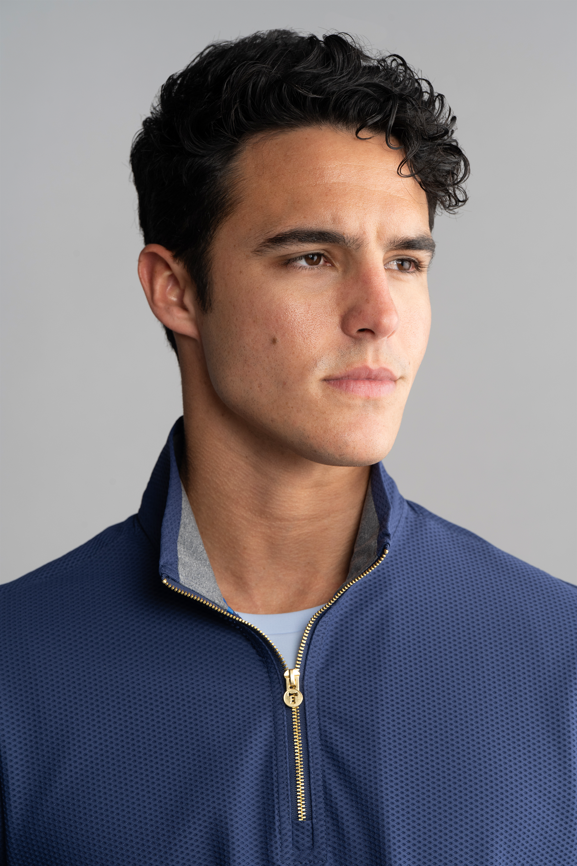 A young man with short, curly dark hair looks right with a neutral expression, wearing the Twillory Air Quarter Zip // Blue. The textured hoodie’s zipper is slightly open over a light shirt, set against a plain light gray background.