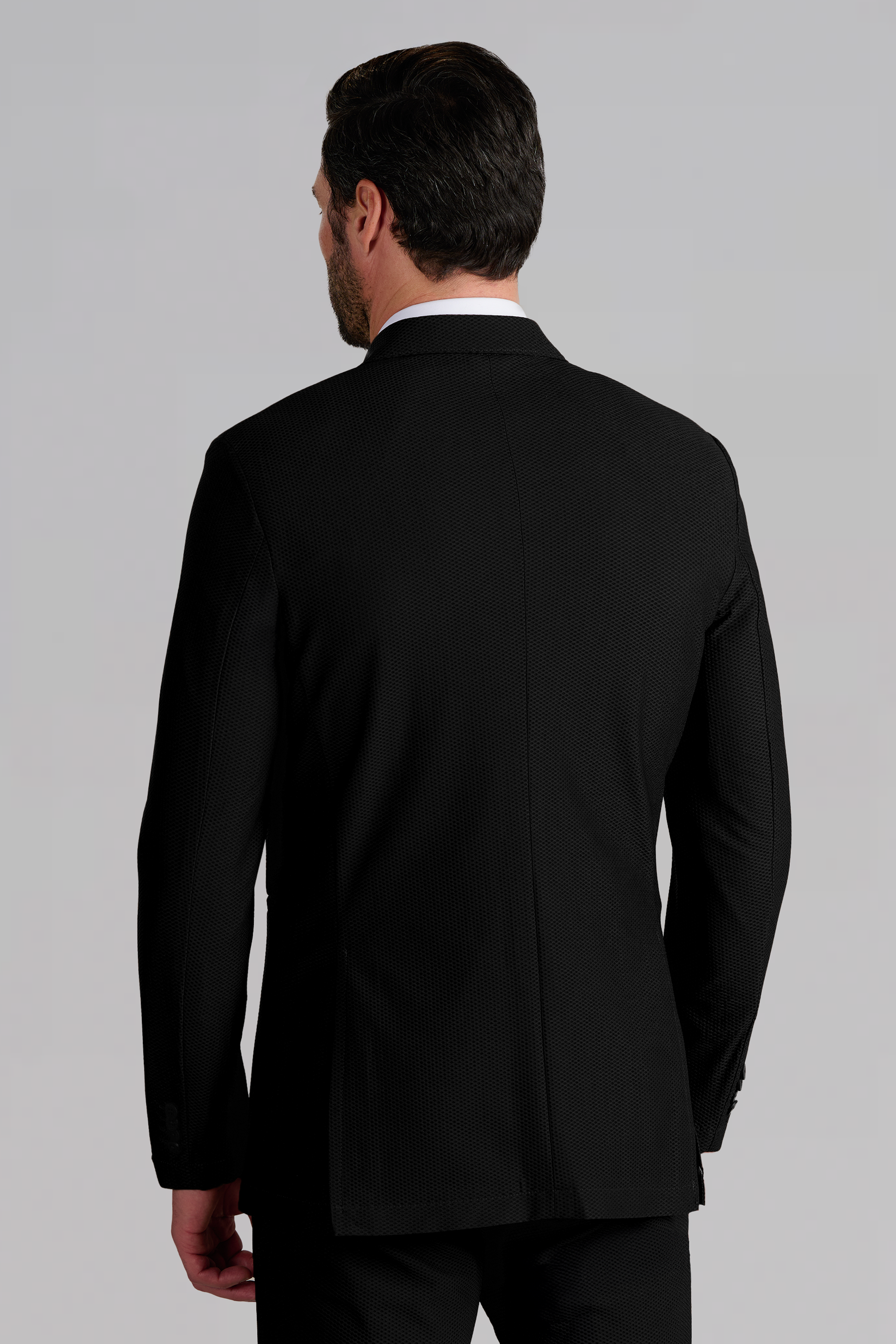 A man with short dark hair is seen from behind, wearing the Twillory AIR Blazer in Black. He stands against a light grey background, left arm slightly bent and right arm relaxed by his side.
