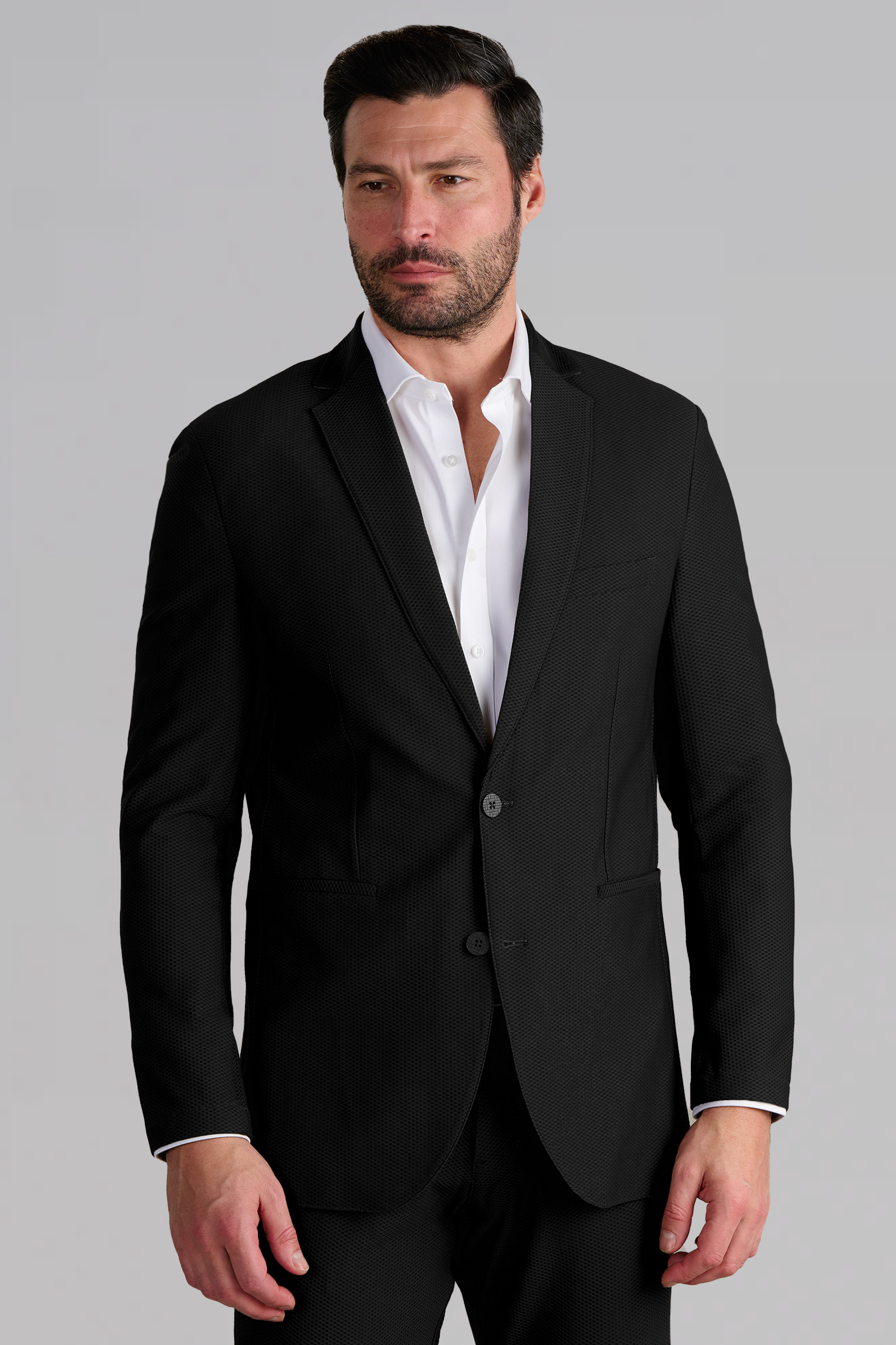 A man with dark hair and a beard wears the Twillory AIR Blazer in black over a white dress shirt. He stands against a plain light gray background, gazing slightly to the side with a neutral expression.